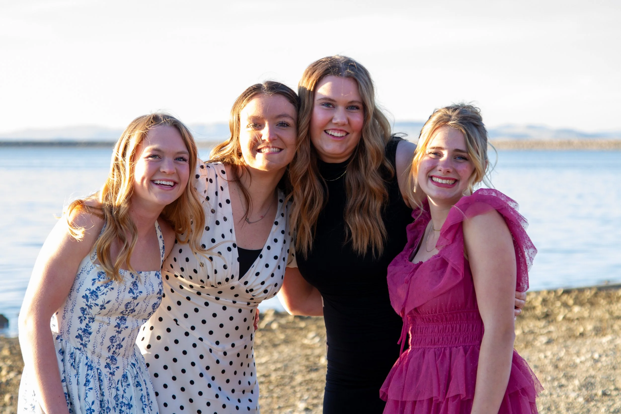 Four young women standing close together on a rocky shoreline near a body of water, smiling at the camera, with a clear sky in the background.