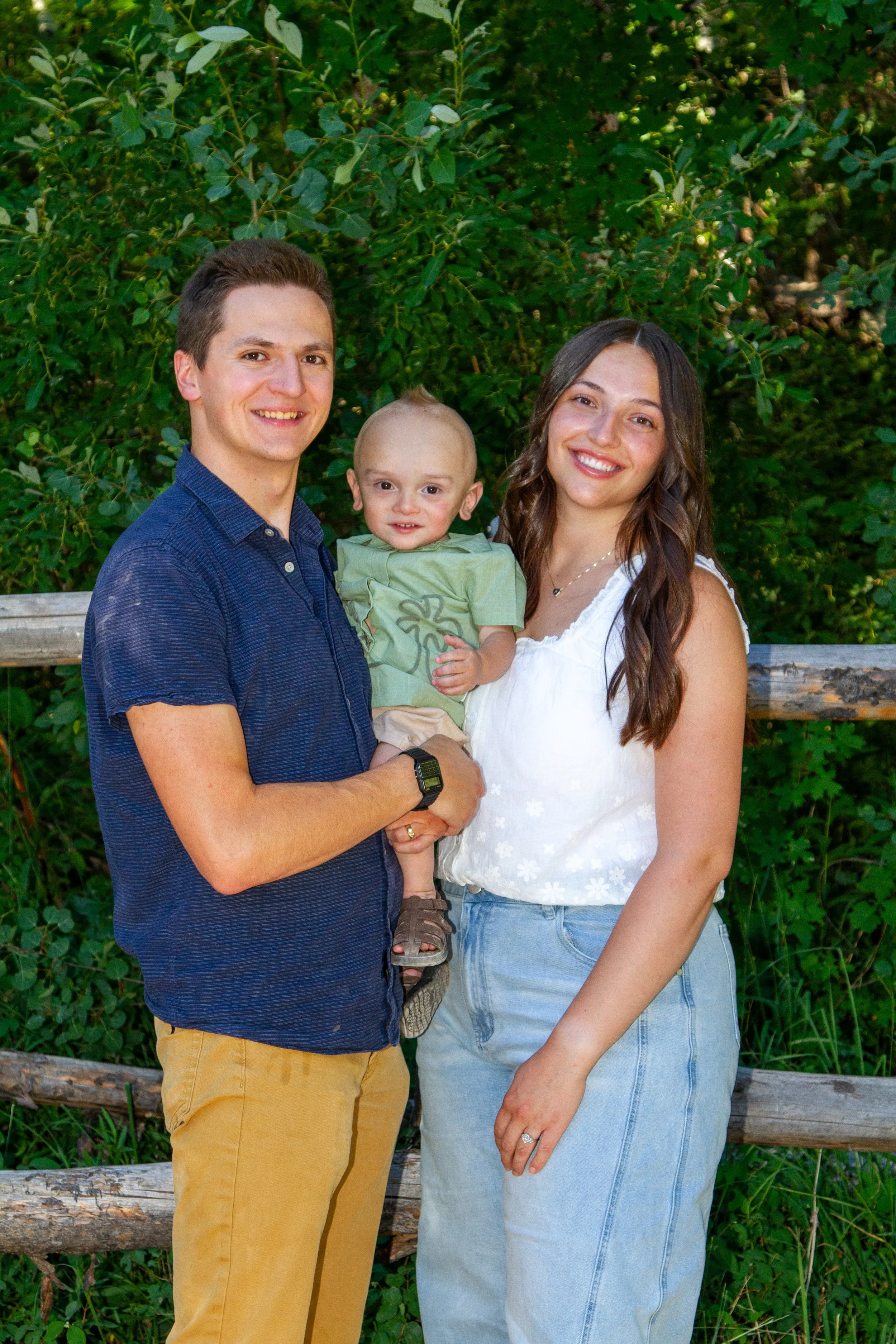 A smiling man and woman stand outdoors in front of green bushes, holding a young child.