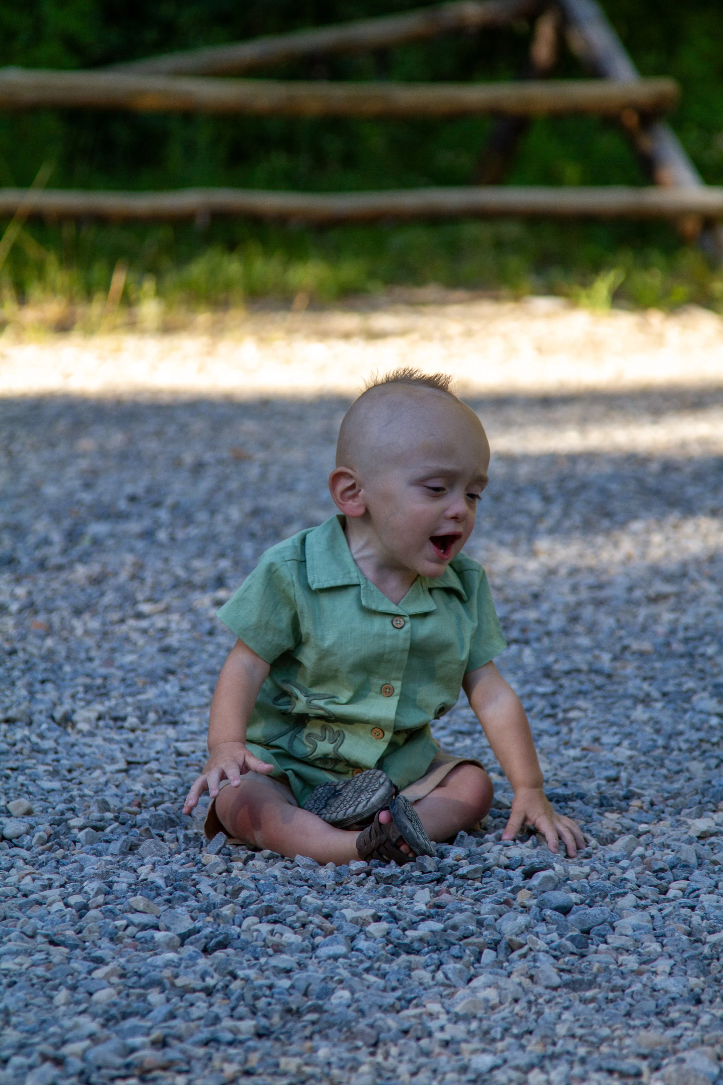 A young child with a shaved head and wearing a green shirt and shorts, sitting on a gravel surface outdoors, with a wooden fence and greenery in the background, appears to be crying or upset.