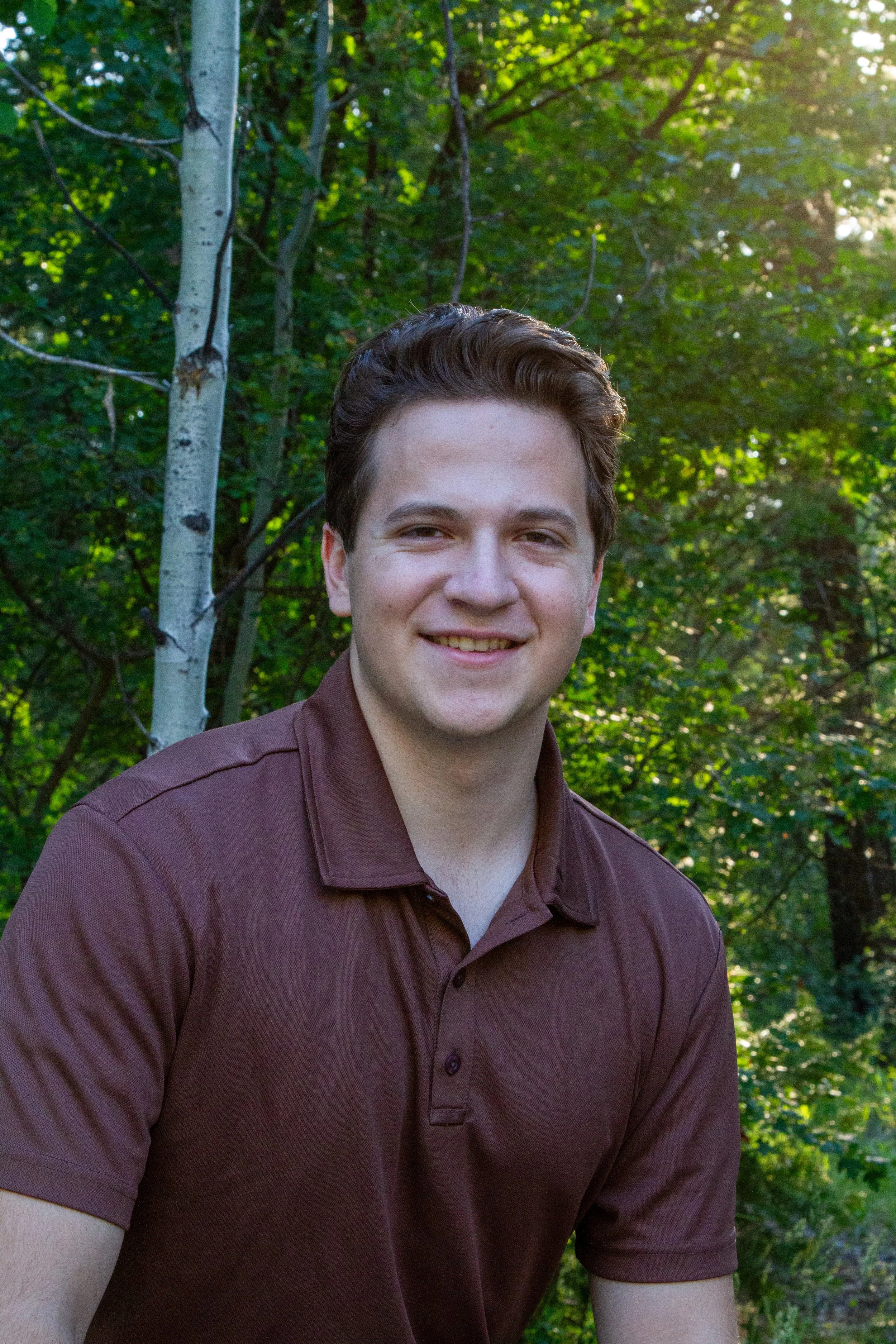 A young man smiling outdoors in front of green trees with sunlight filtering through.