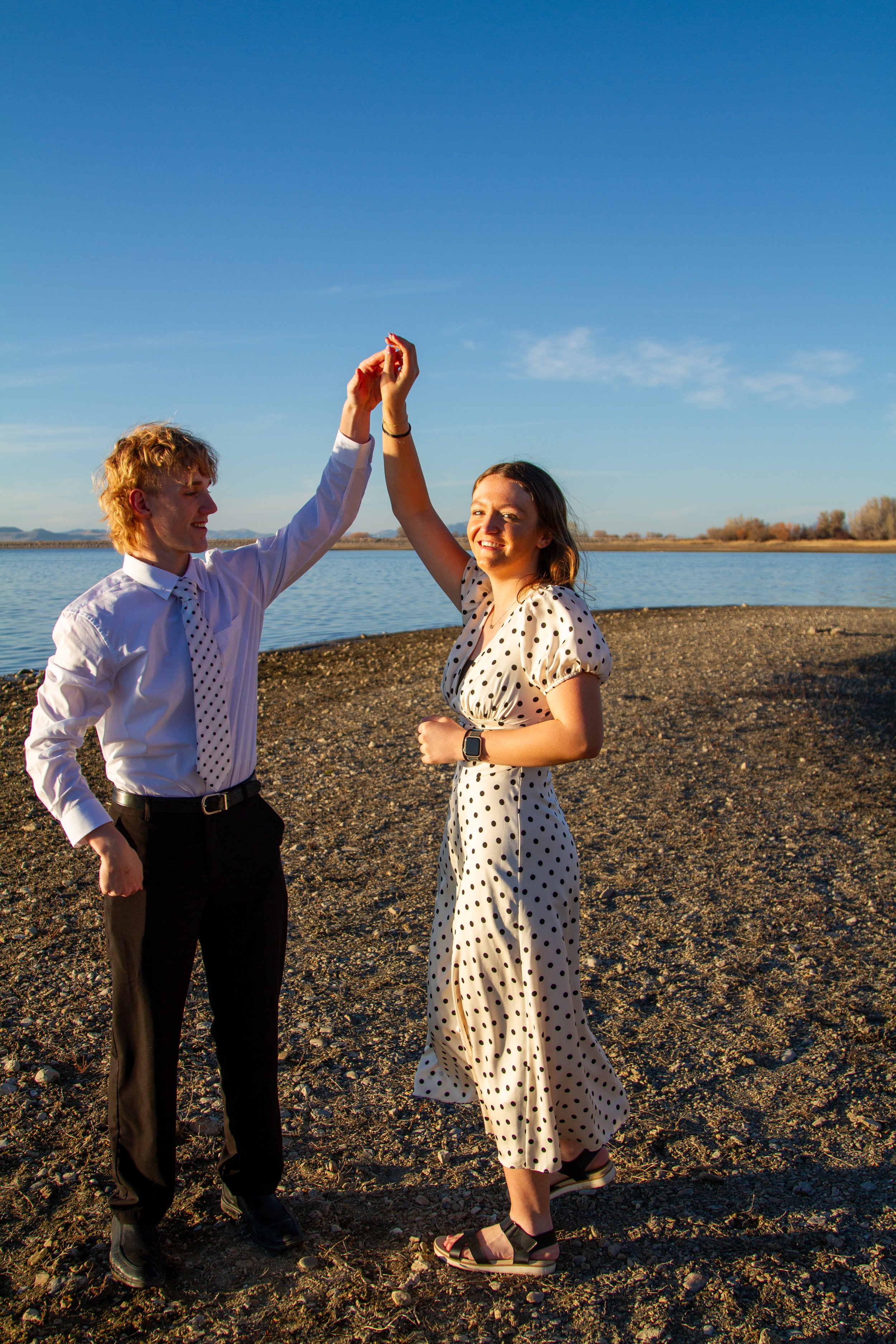 A young couple dancing hand-in-hand on a rocky shoreline with a lake and clear blue sky in the background.