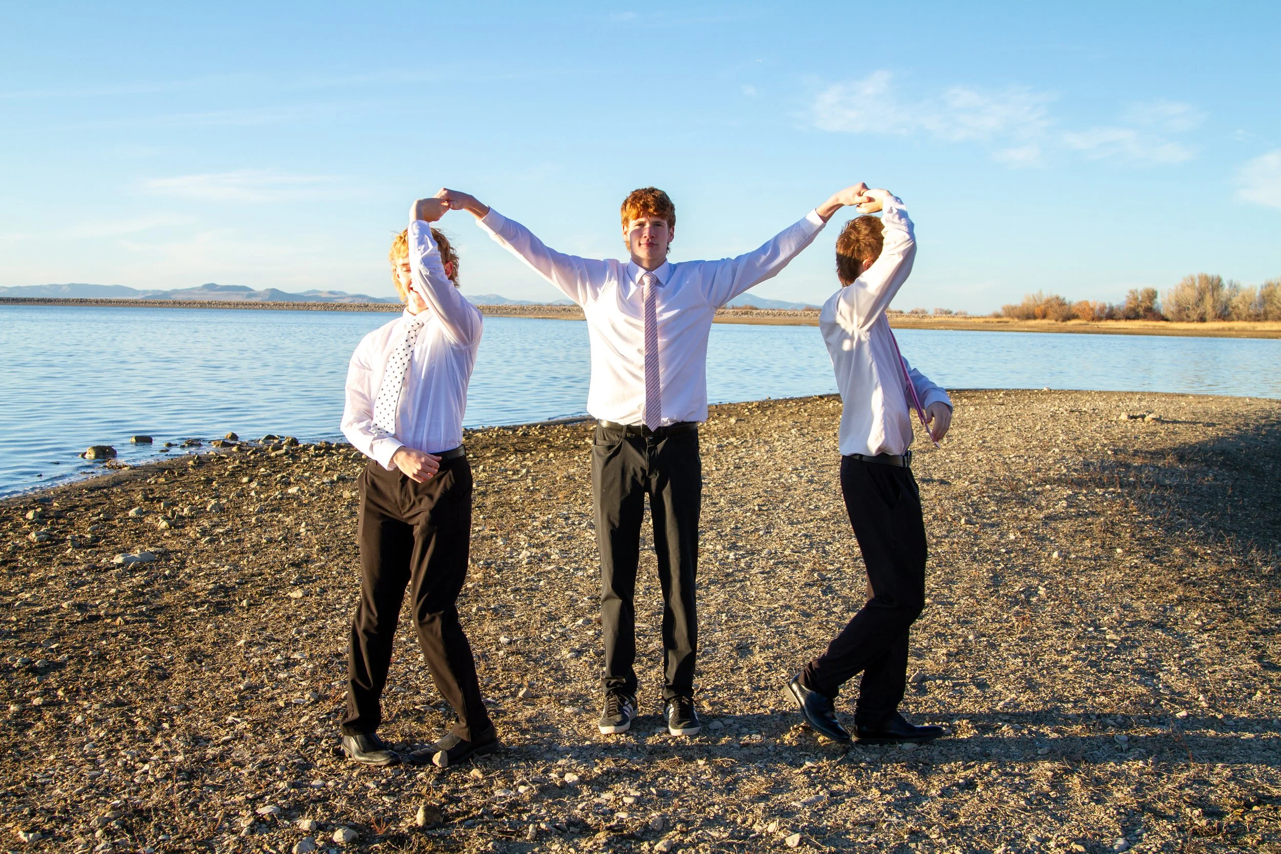 Three young men in business attire standing on a rocky shoreline near a body of water, holding hands above their heads forming an arc, with the middle person raising both arms, in a sunny outdoor setting.