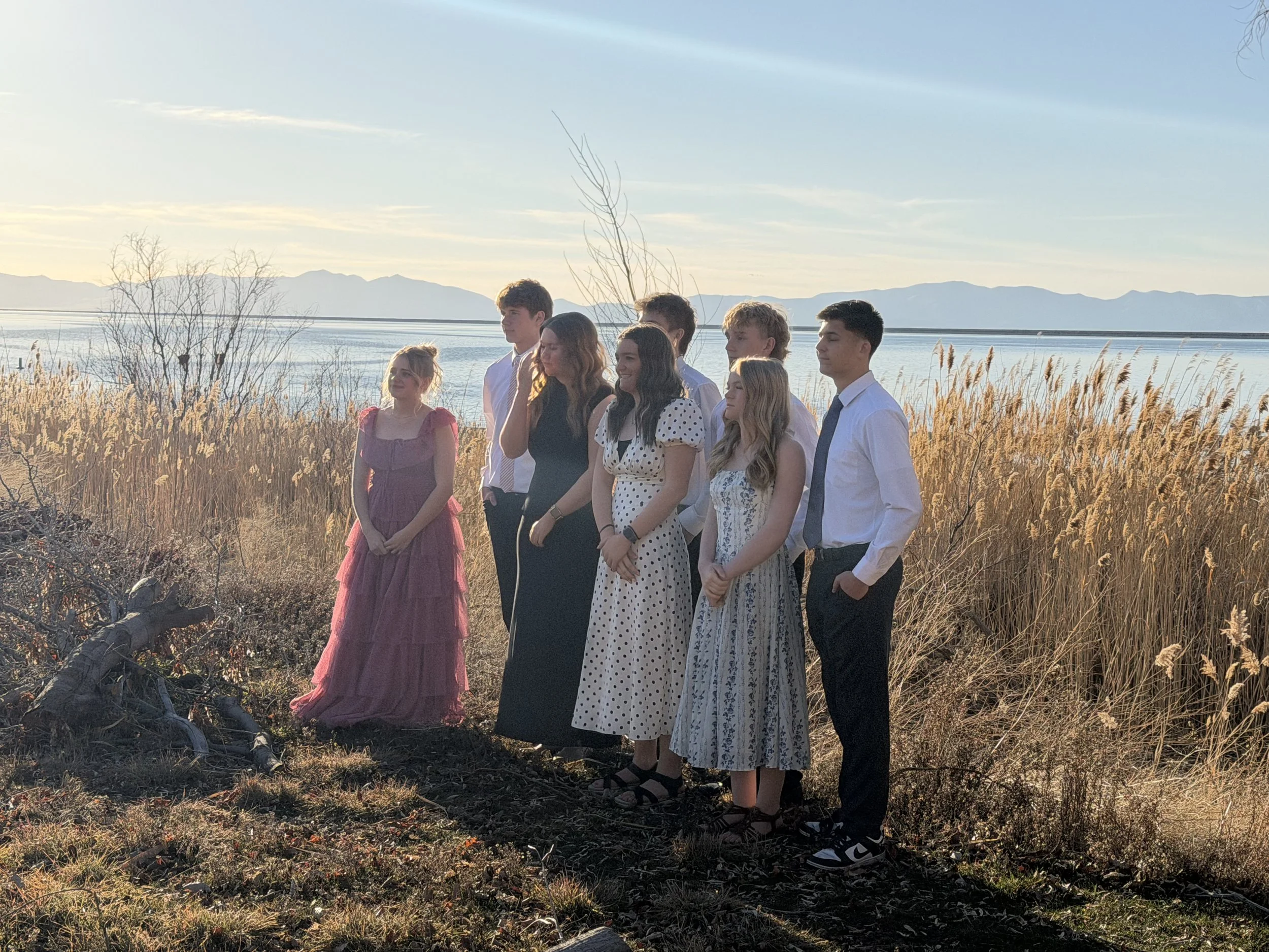 A group of eight young people dressed in semi-formal attire standing outdoors near a lake with mountains in the background, during sunset.