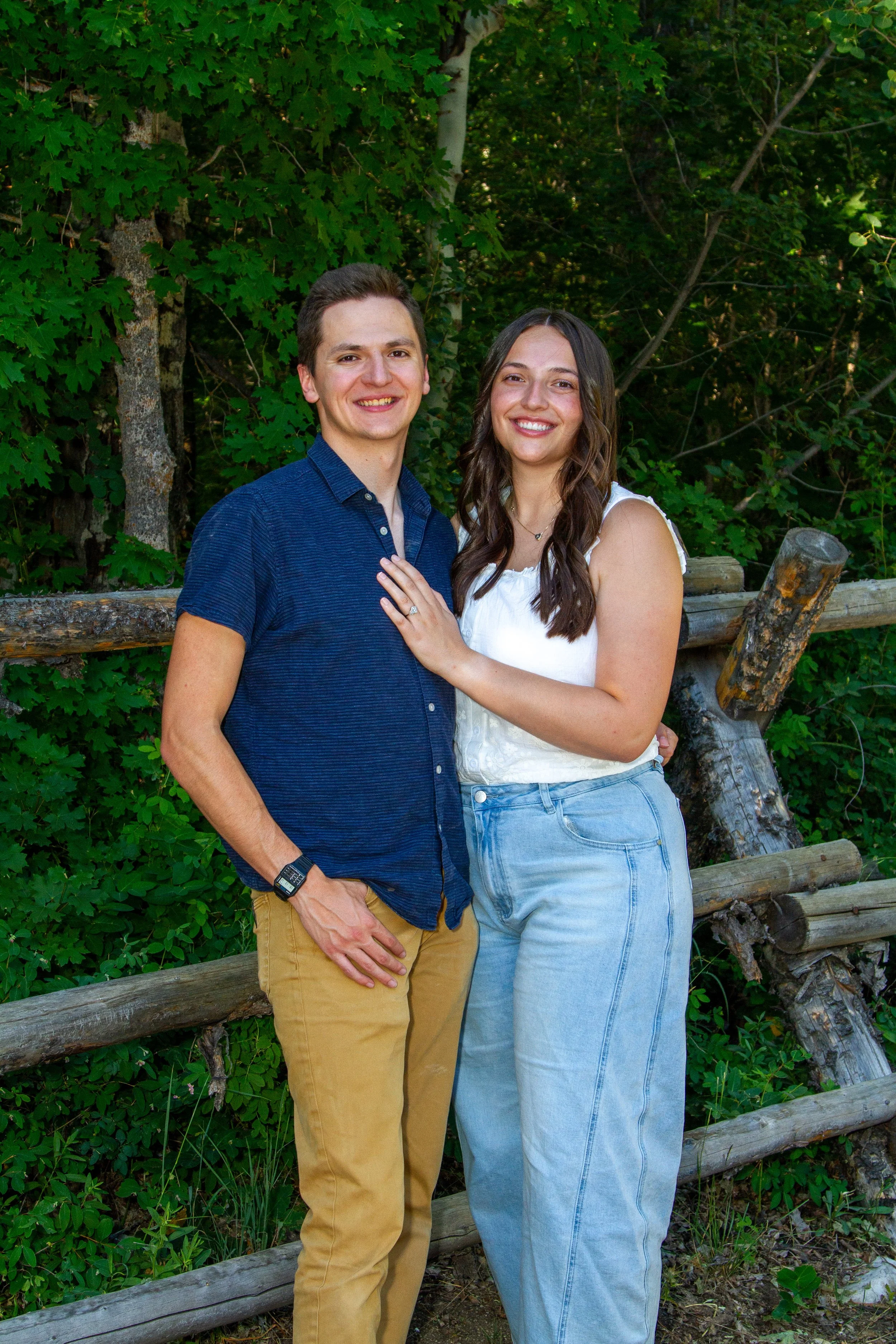 A smiling young man and woman standing close together outdoors with a green leafy background and a wooden fence.