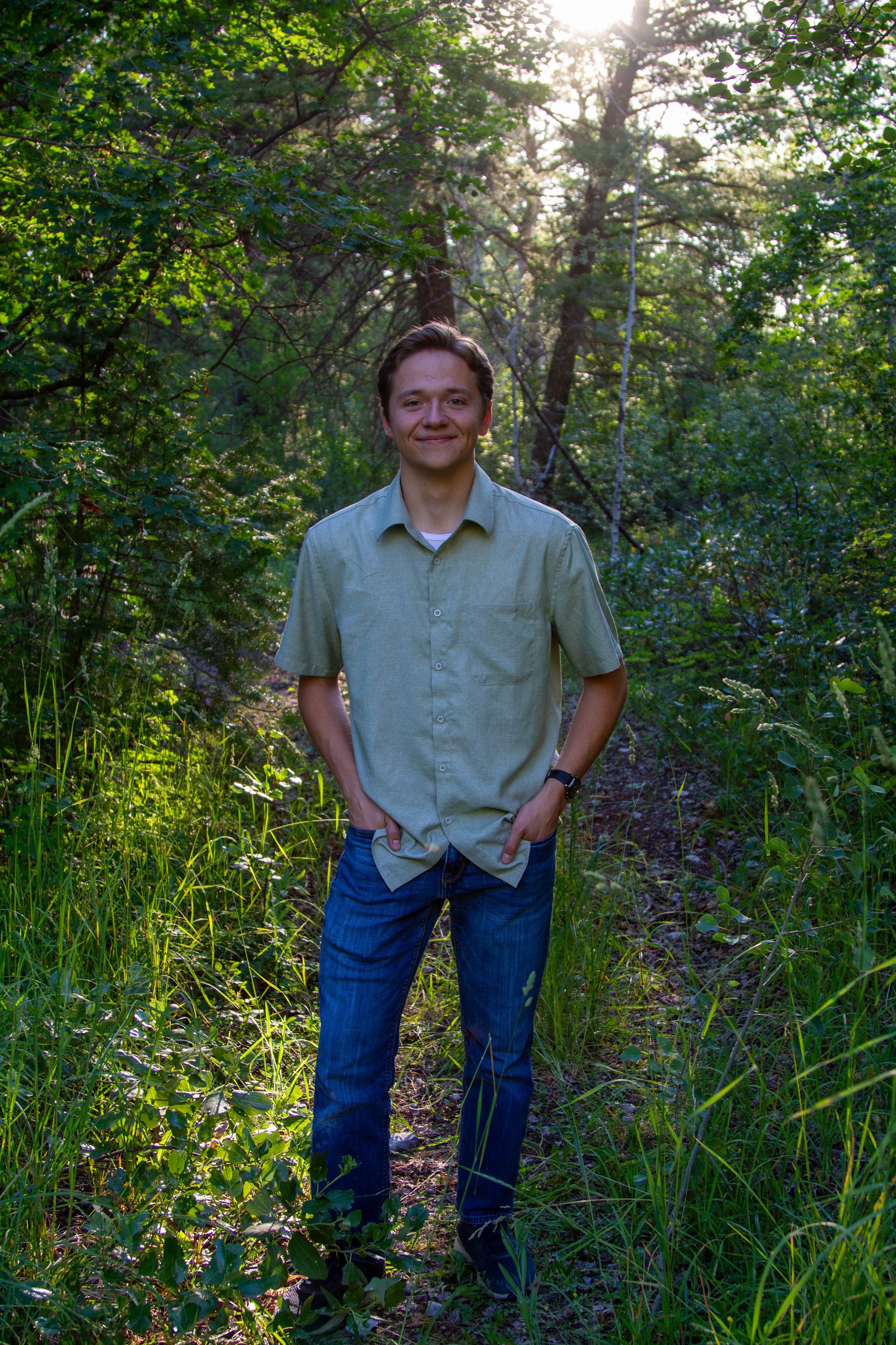 A young man standing on a forest trail, wearing a light green button-up shirt and dark blue jeans, with tall green trees and sunlight in the background.