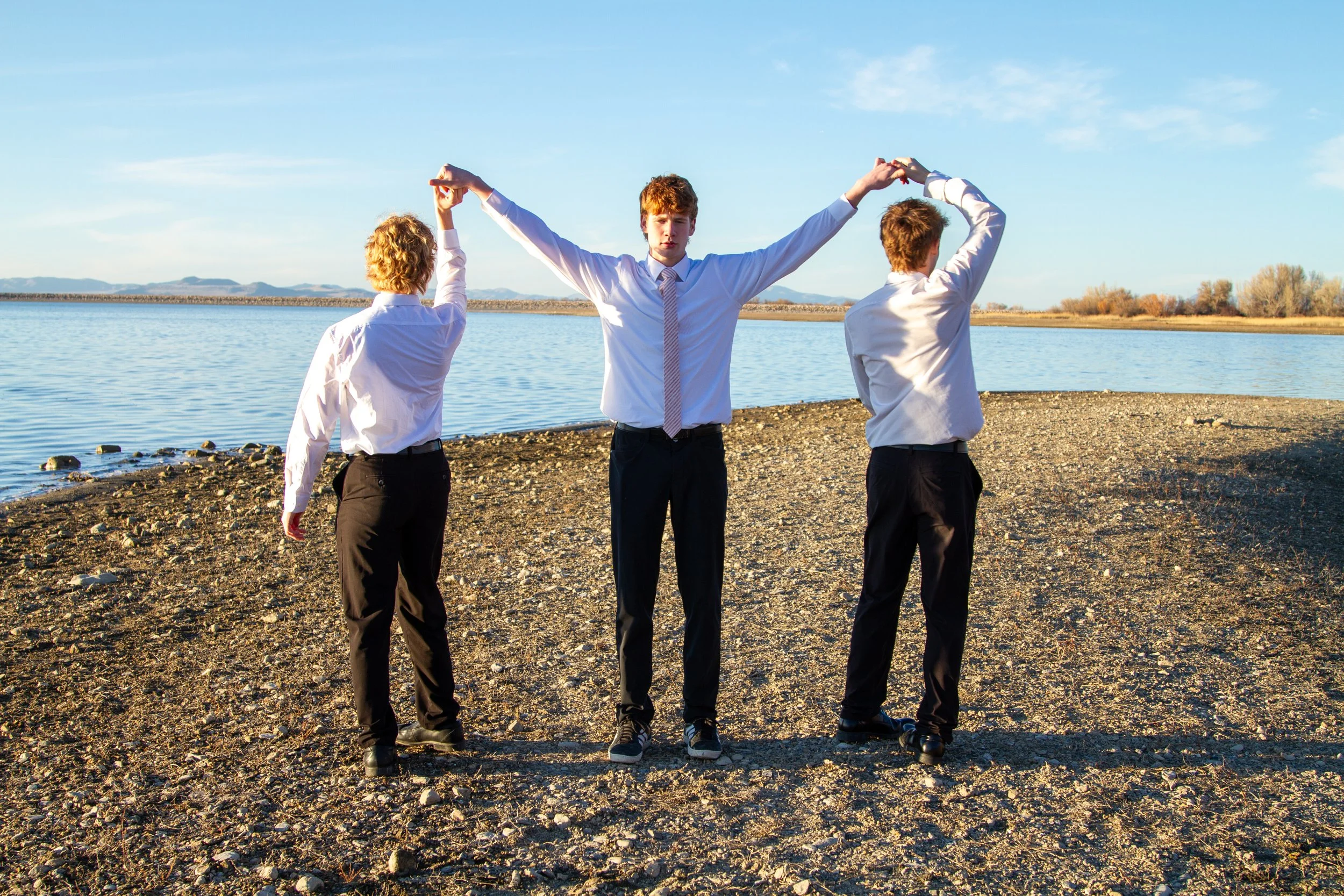 Three young men dressed in white shirts and black pants standing on a rocky shoreline by a body of water, holding hands and raising their arms together under a blue sky.