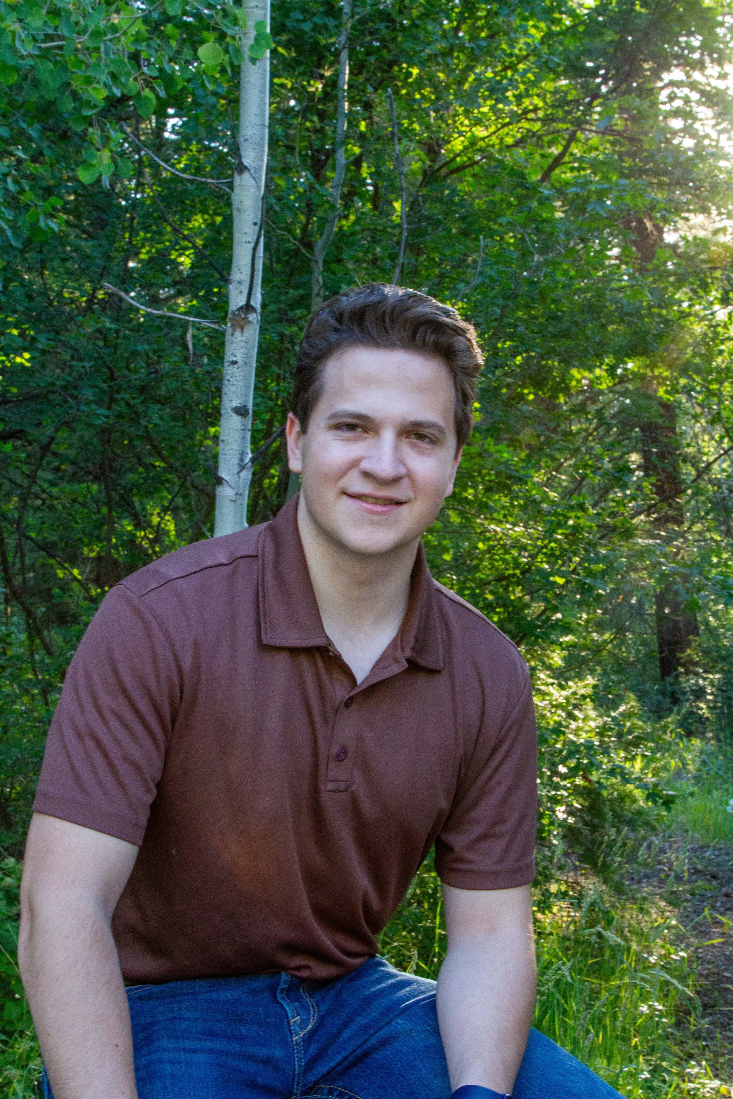 A young man with dark hair wearing a brown polo shirt and blue jeans sitting outdoors in a lush green forested area with sunlight filtering through the leaves.