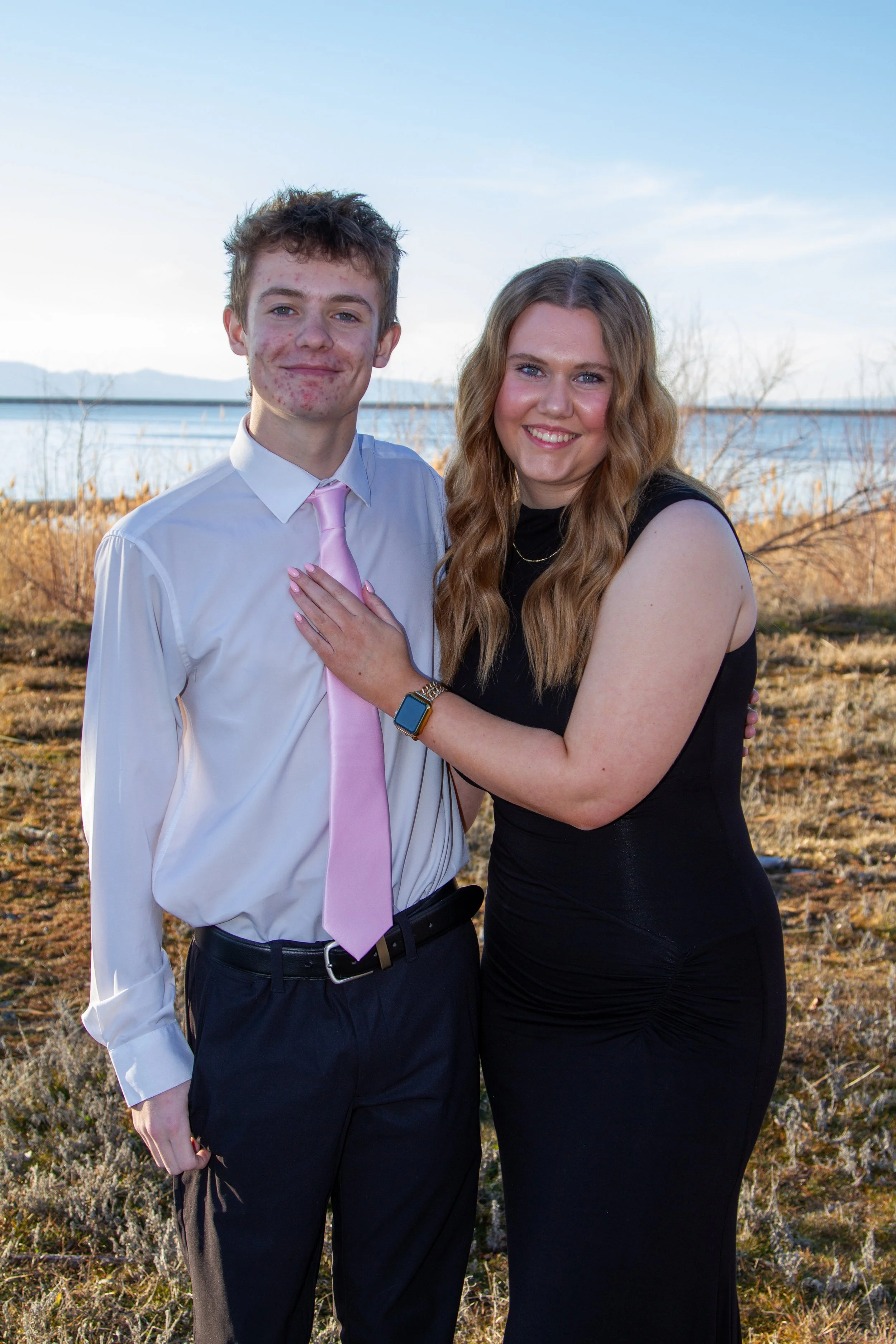 A young man and woman standing outdoors by a body of water, smiling at the camera. The young man is wearing a white dress shirt, pink tie, and dark dress pants. The woman is wearing a sleeveless black dress and has long, wavy, blonde hair.