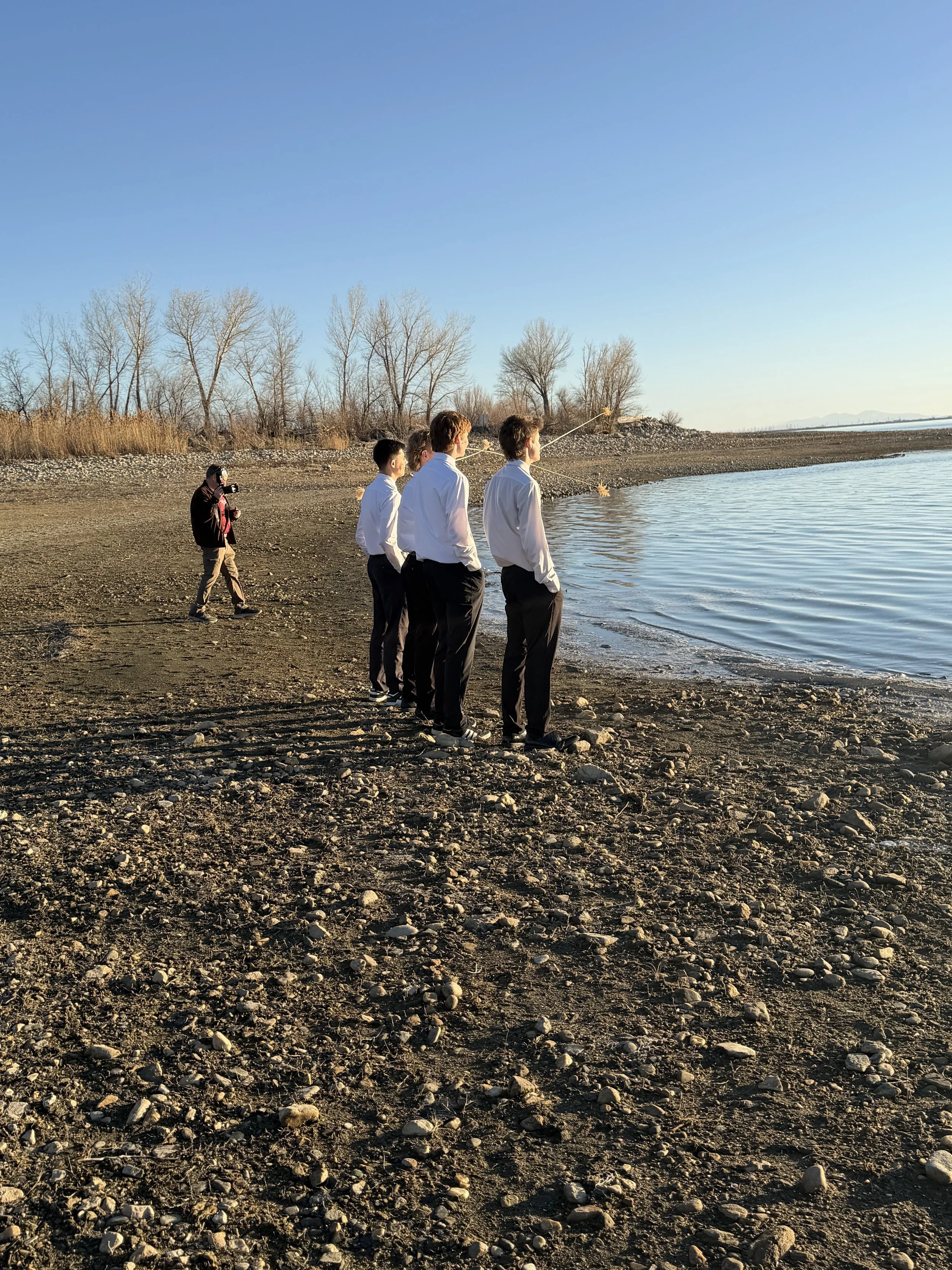 Four people in white shirts and black pants stand by a lakeshore, fishing, with a person in black and brown walking nearby. Bare trees are in the background under a clear blue sky.