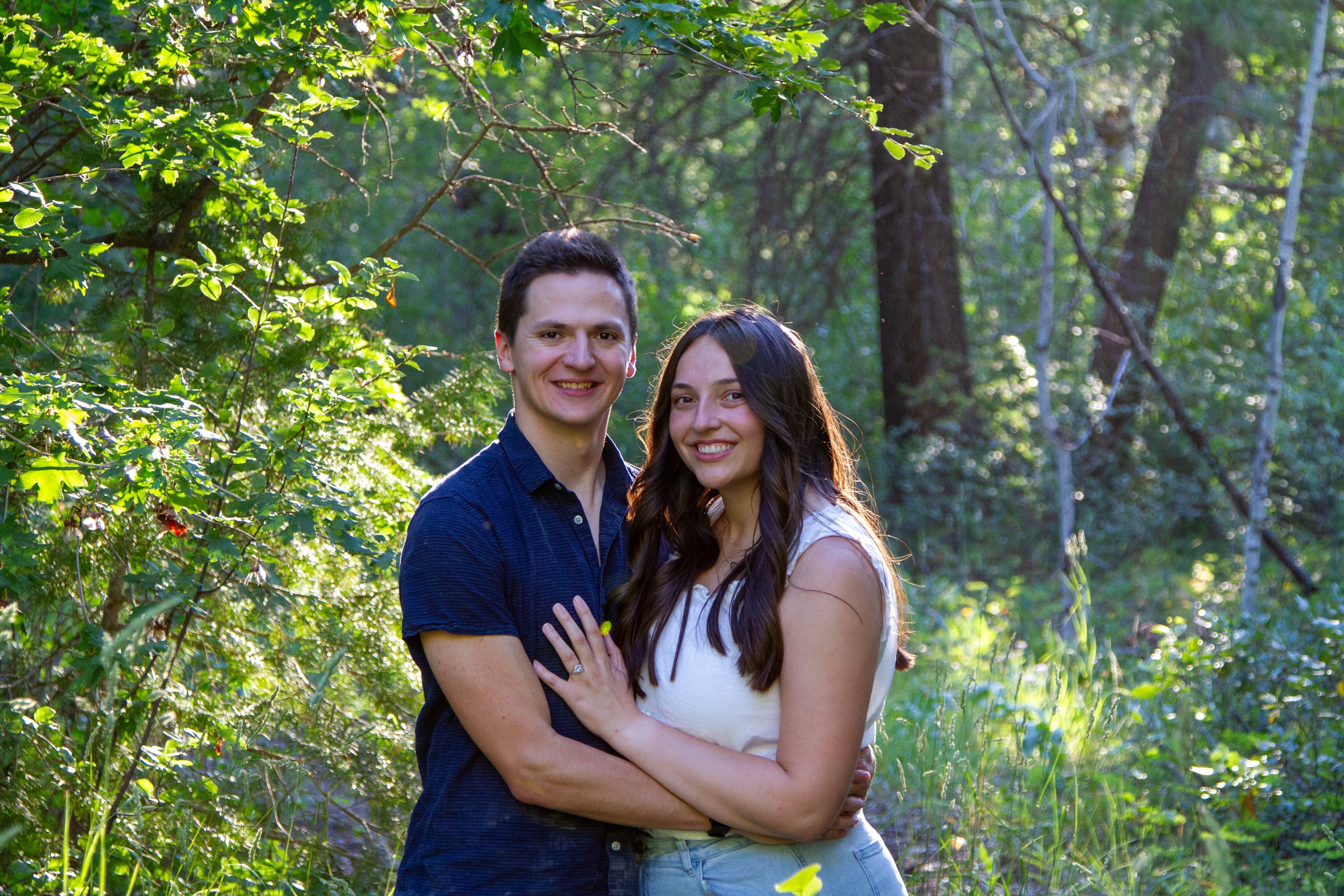 A young couple hugging and smiling in a forest with green foliage and sunlight filtering through the trees.