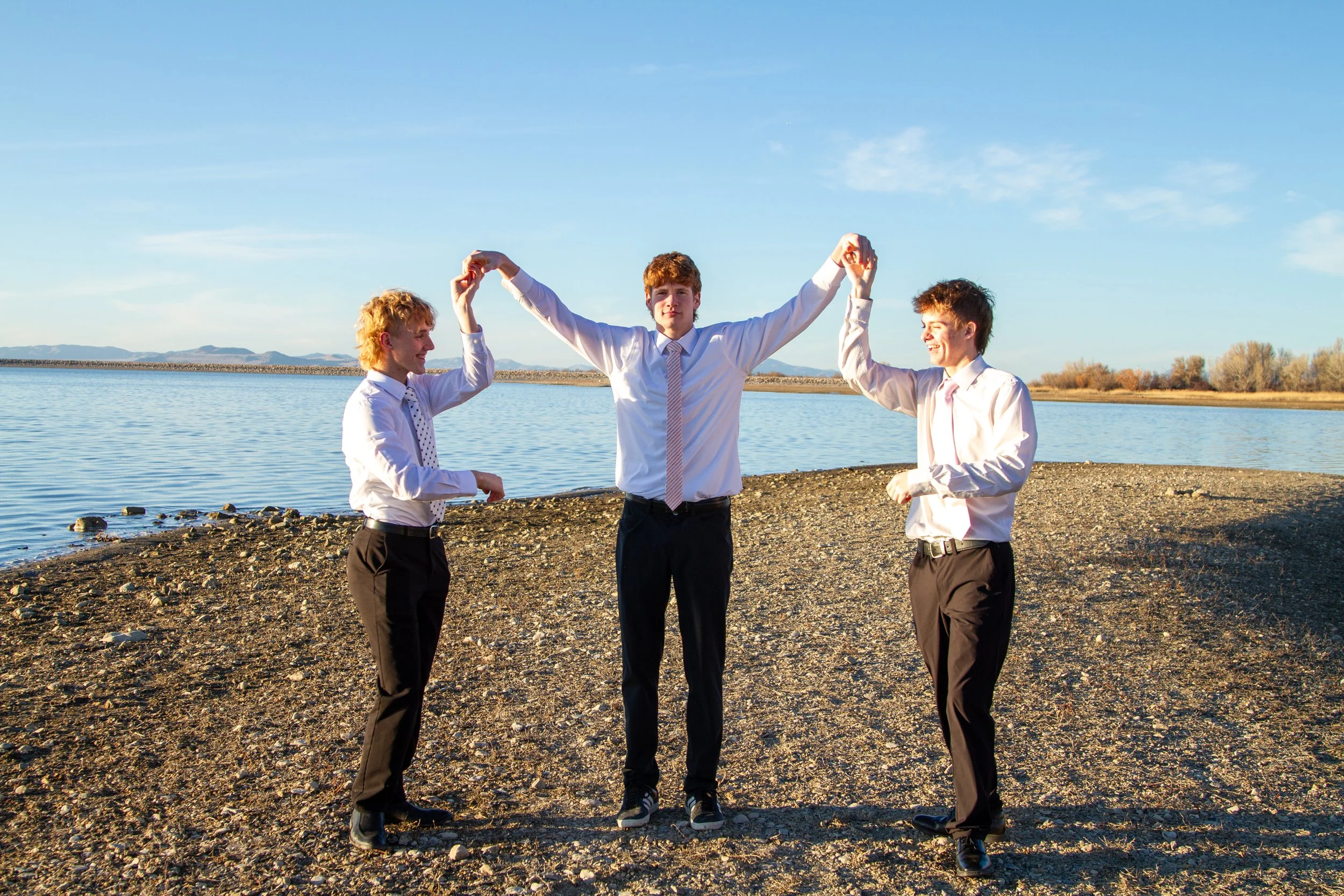 Three young men in dress shirts and trousers standing on a rocky shoreline by a body of water, holding hands and celebrating, with mountains and trees in the background on a sunny day.