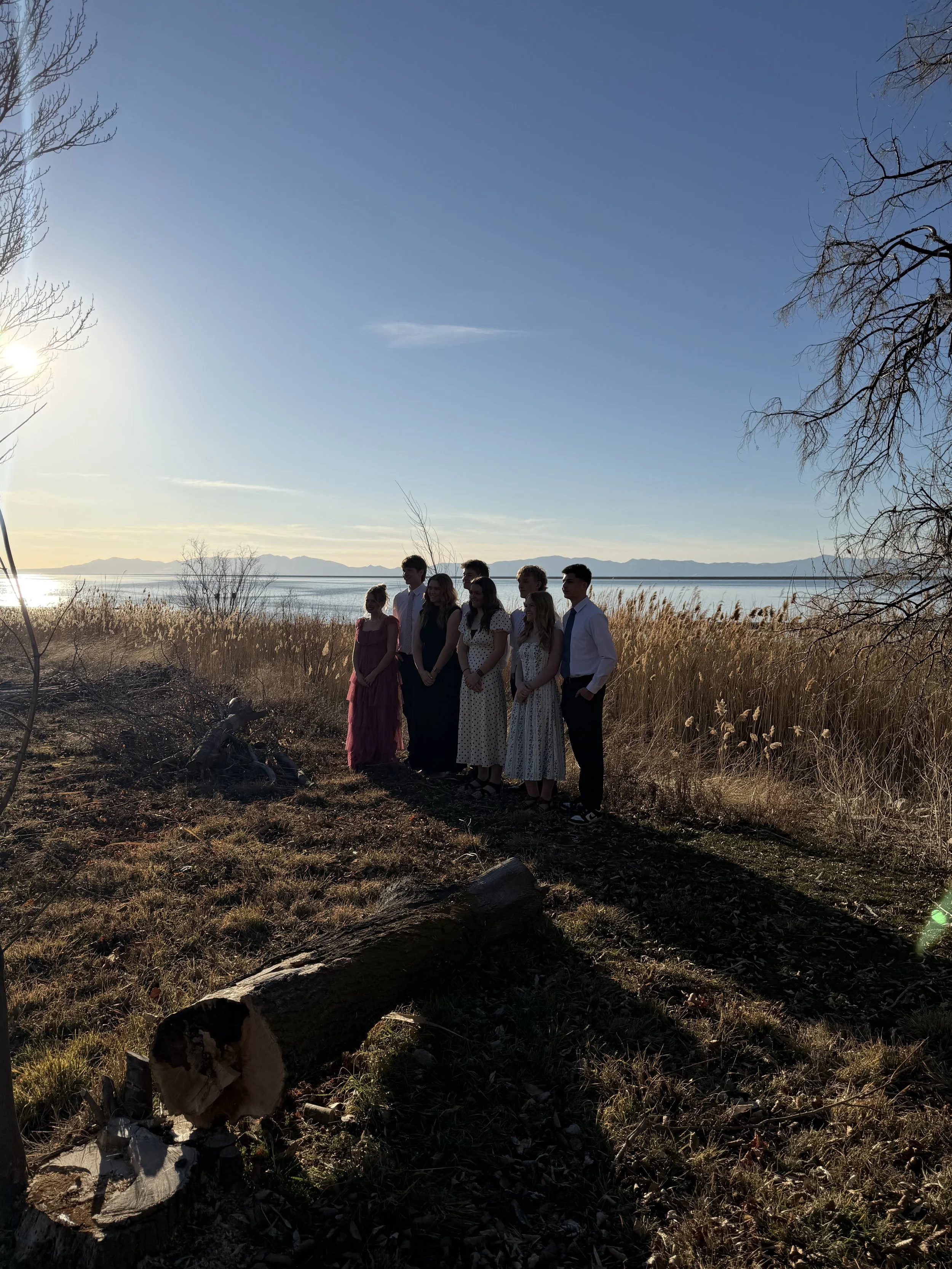 A group of six people standing outdoors near a body of water during sunset, with tall grass and trees around them.