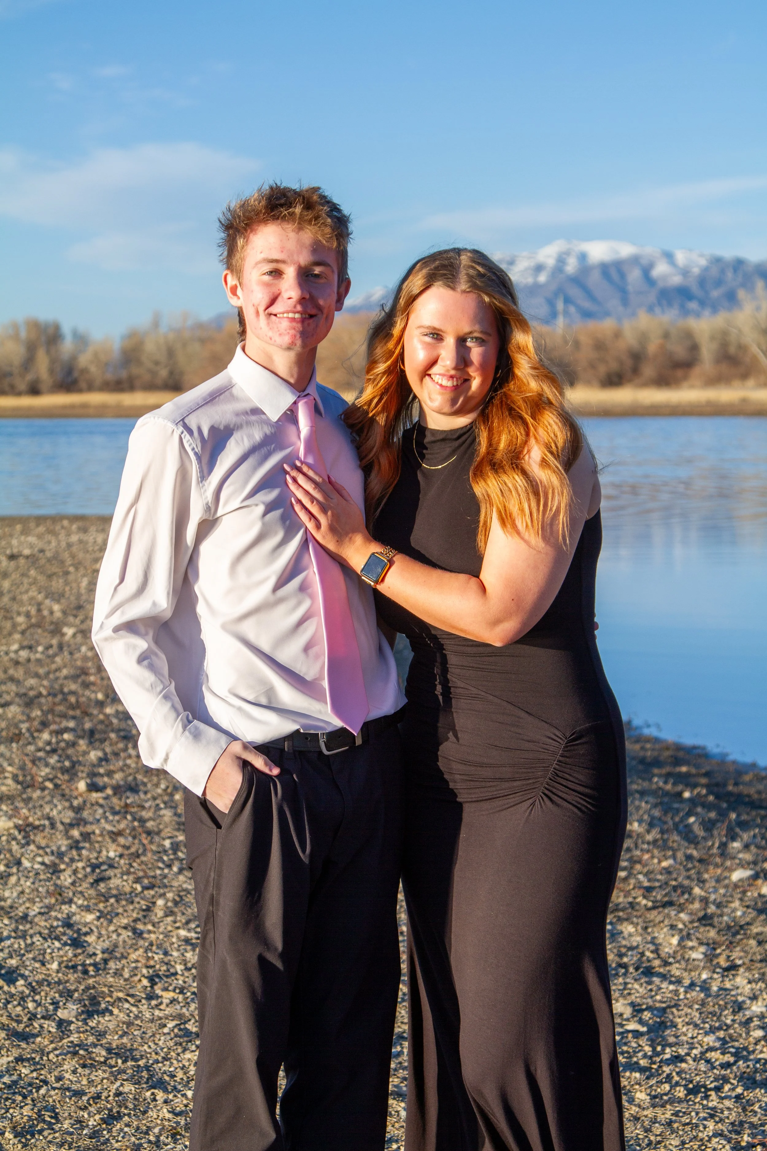 A young couple standing by a river with mountains in the background, smiling at the camera.