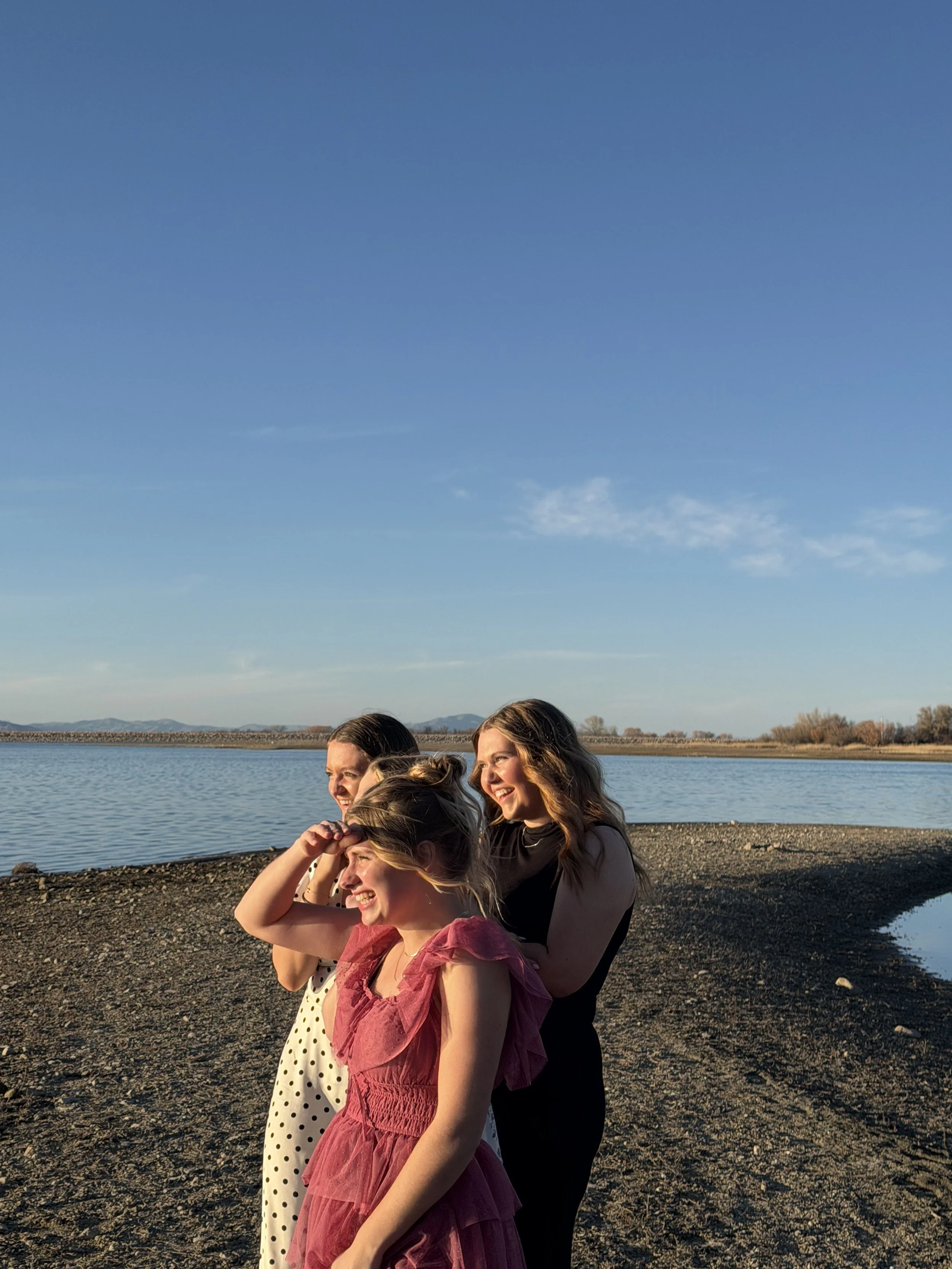 Three women standing on a pebbled shoreline near a large body of water, with mountains in the distance, enjoying sunny weather and smiling.
