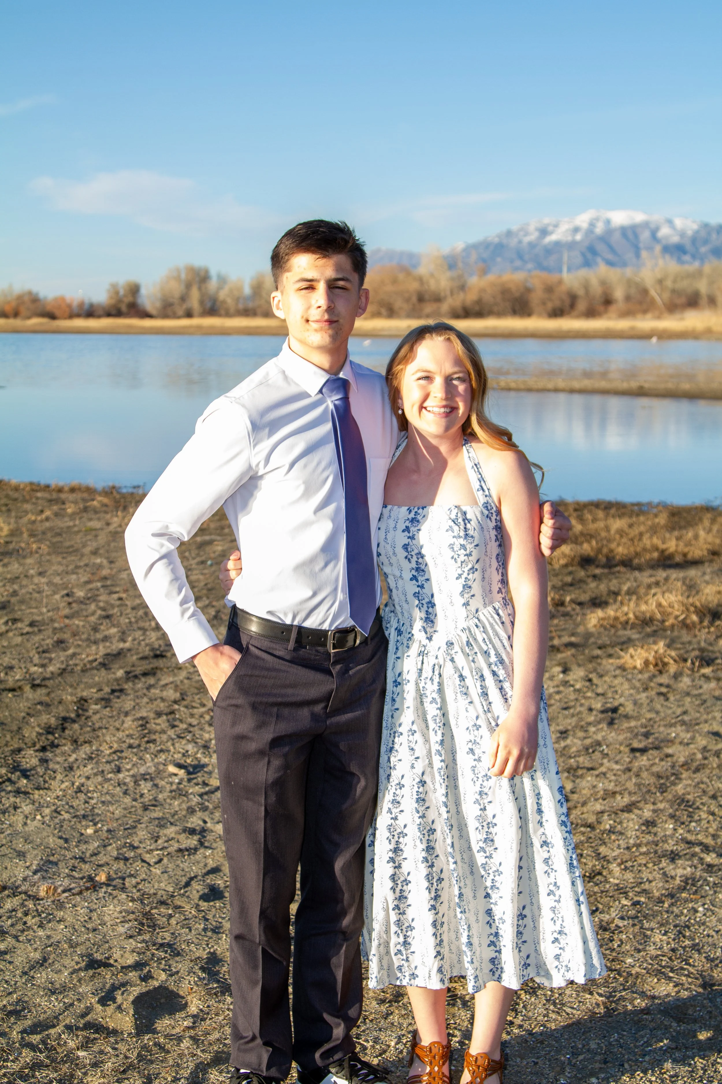 A young man in a white shirt and dark trousers standing next to a young woman in a white and blue floral dress, smiling, with a body of water and mountains in the background.