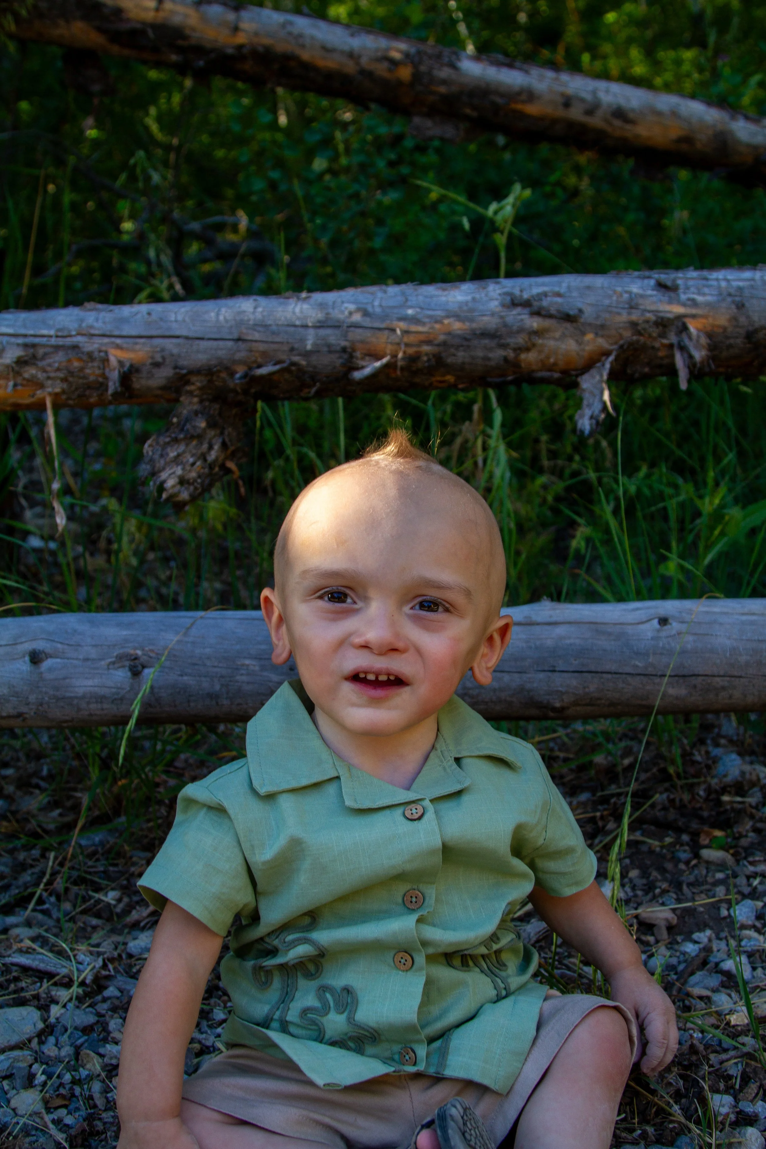 A young boy with light skin and short blonde hair sitting on the ground outdoors, smiling, with a wooden fence and green foliage in the background.