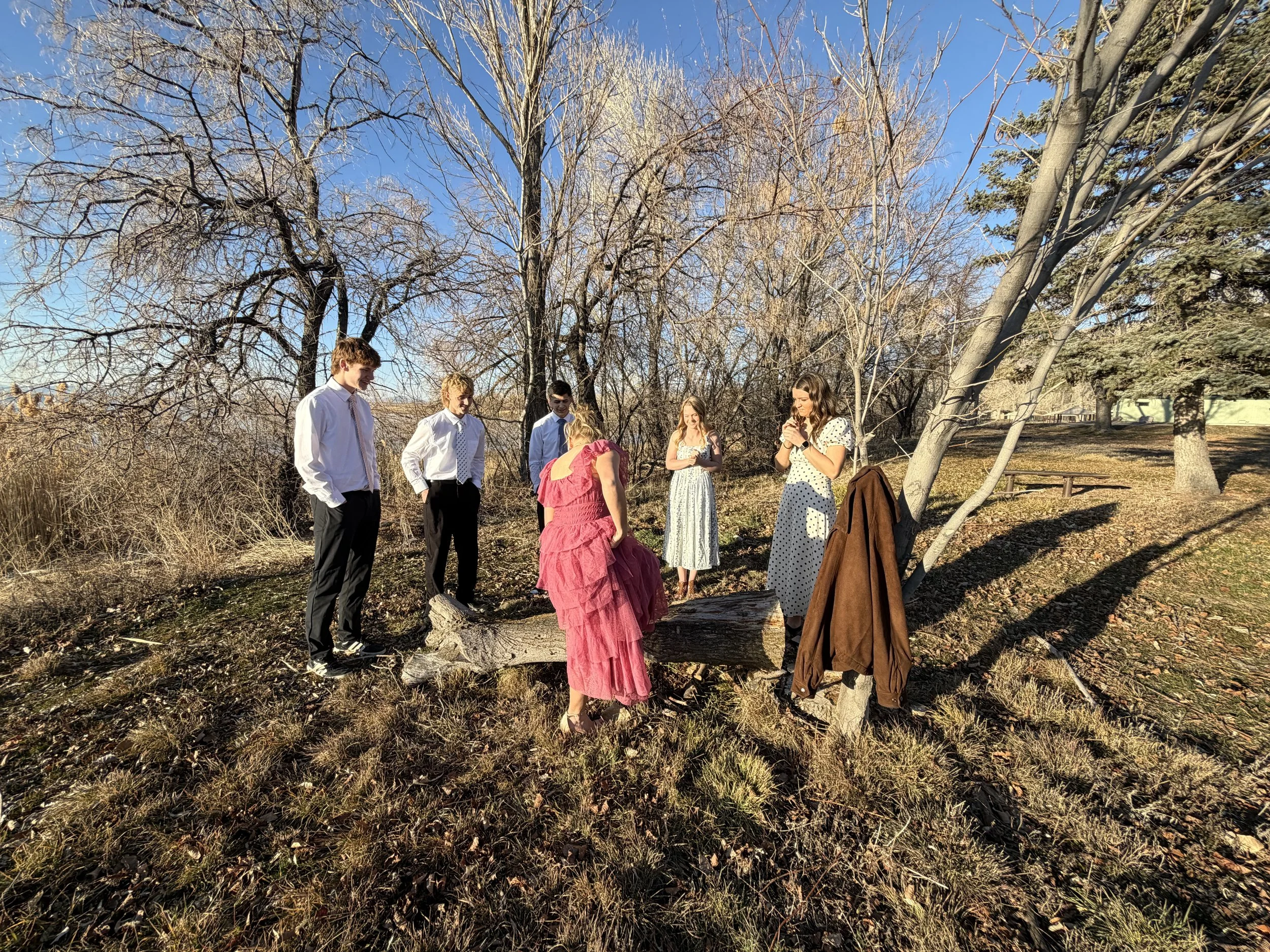 People dressed for a wedding ceremony outdoors, with one woman in a pink dress standing on a fallen log and others in formal attire standing around her, under leafless trees on a sunny day.