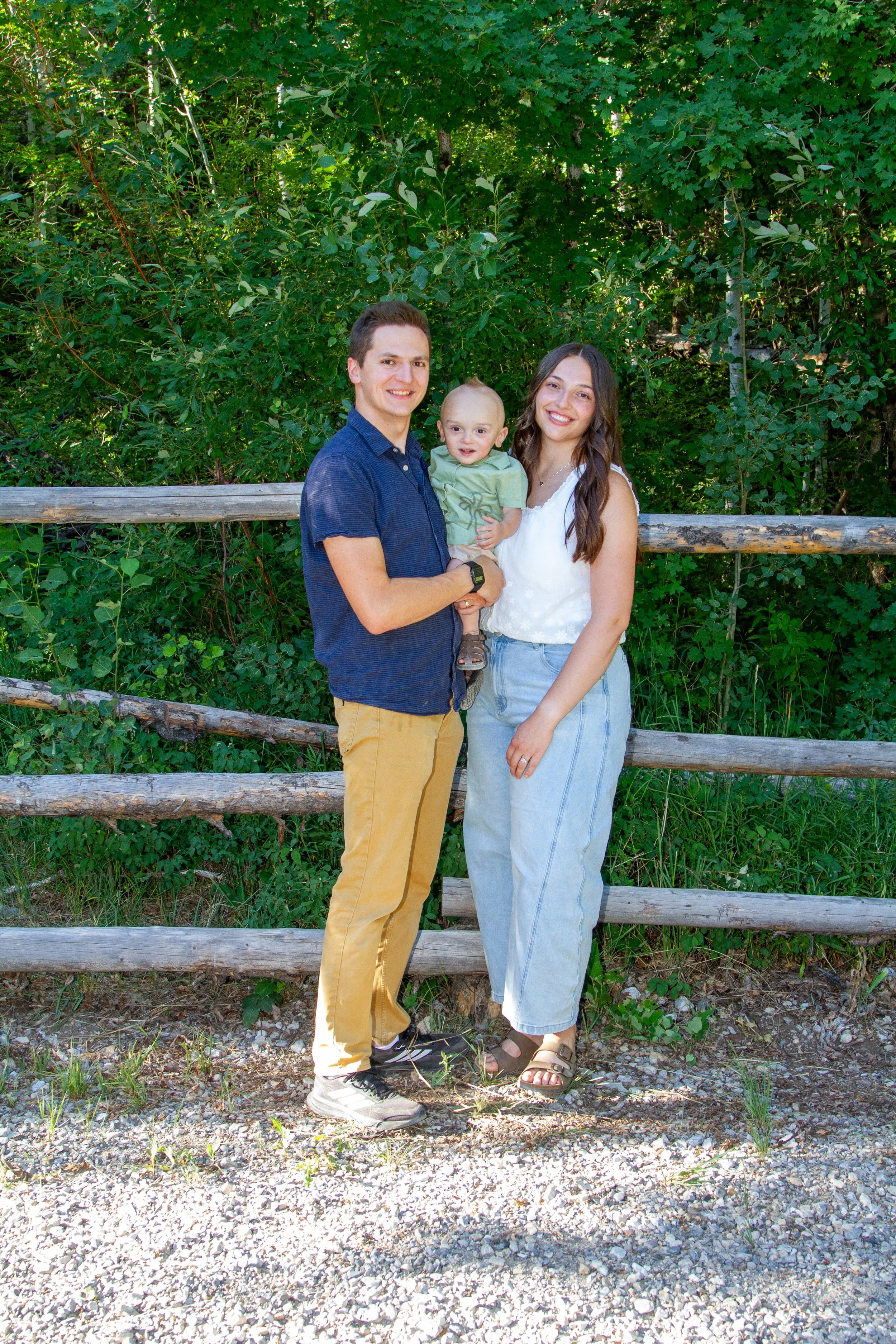 A family of three standing outdoors next to a wooden fence, surrounded by green trees. The man is holding a young child, and the woman stands beside them, all smiling at the camera.