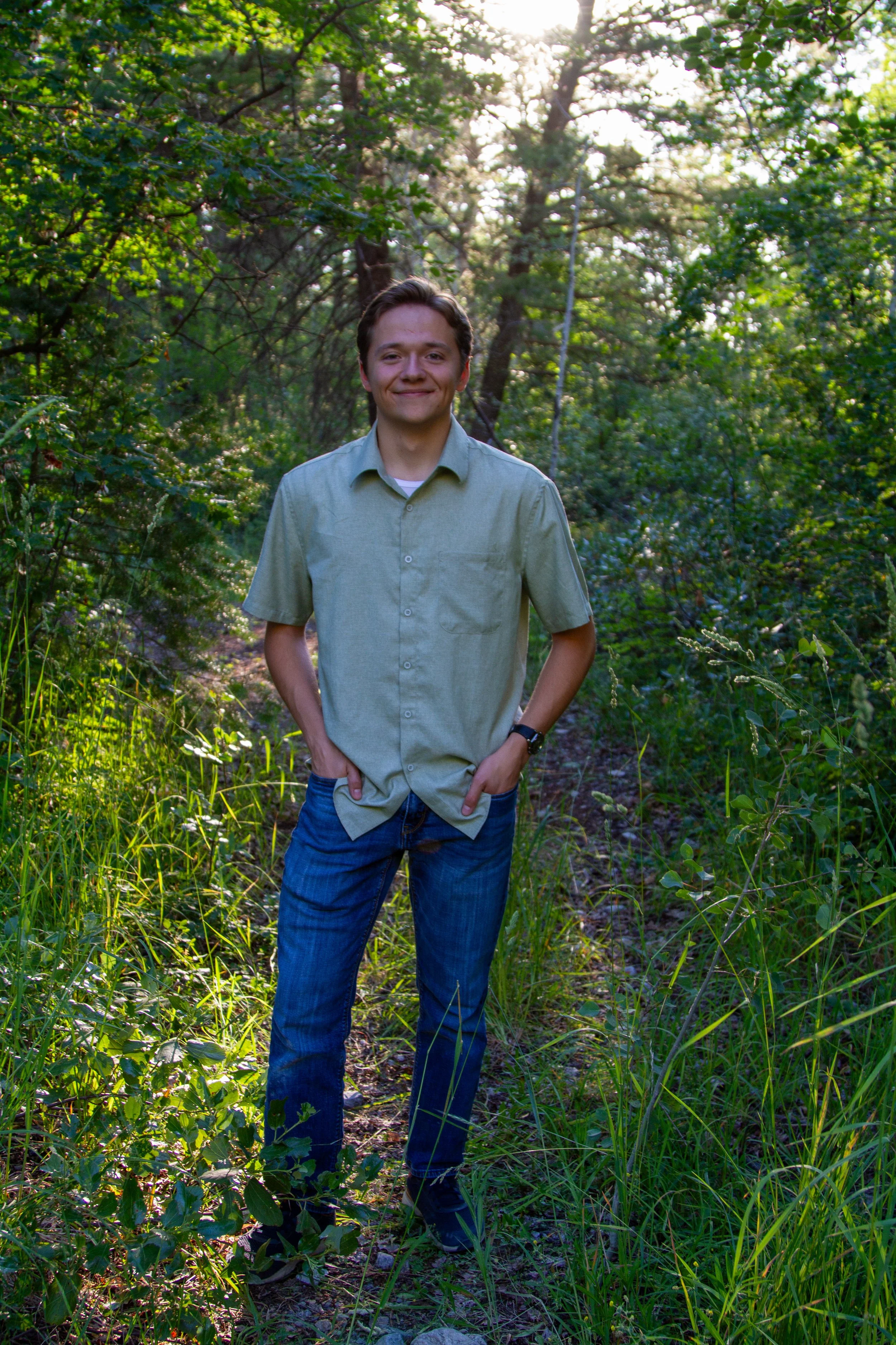 A young man in a green shirt and jeans standing on a narrow dirt trail surrounded by green trees and tall grass in a forest during daylight.