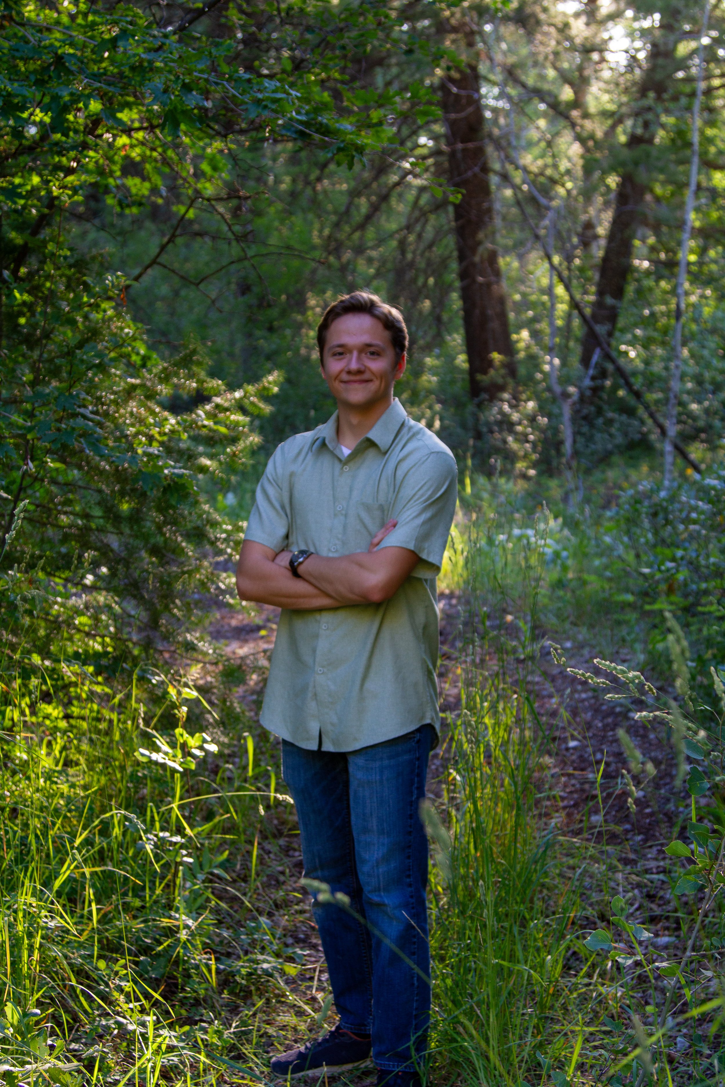 A young man in a light green button-up shirt and blue jeans standing outdoors in a forest with arms crossed, surrounded by green foliage and trees, during daylight hours.