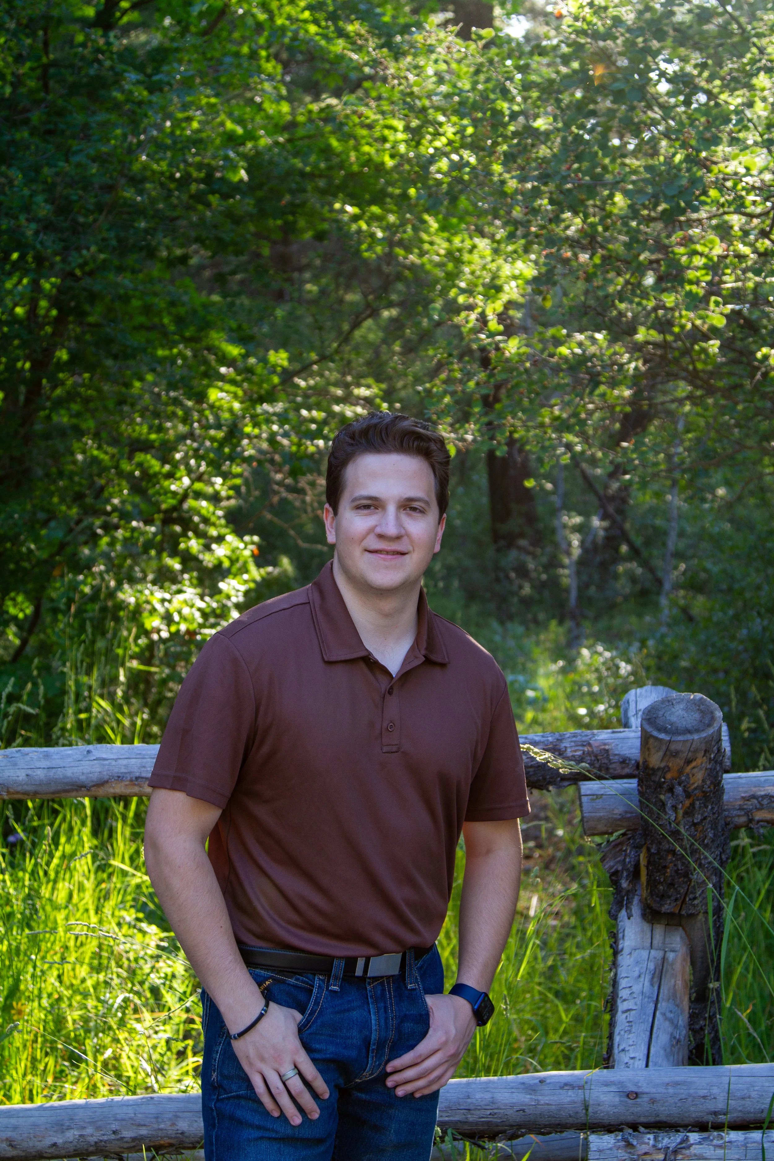A young man with dark hair smiling, wearing a brown polo shirt, blue jeans, and a black watch, standing outdoors near a wooden fence with lush green trees and grass in the background.