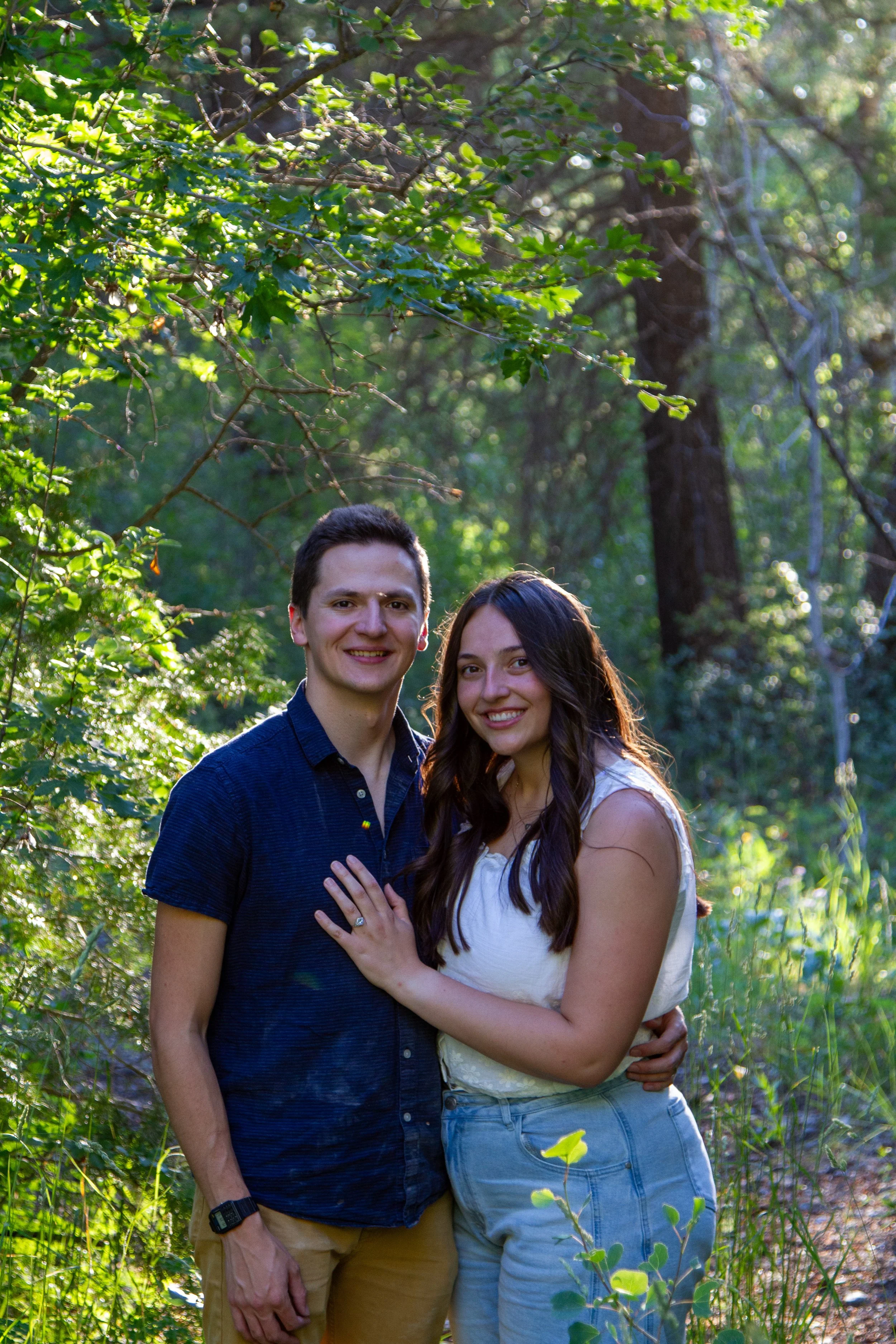 A smiling man and woman standing close together in a lush, green forest, with their arms around each other.