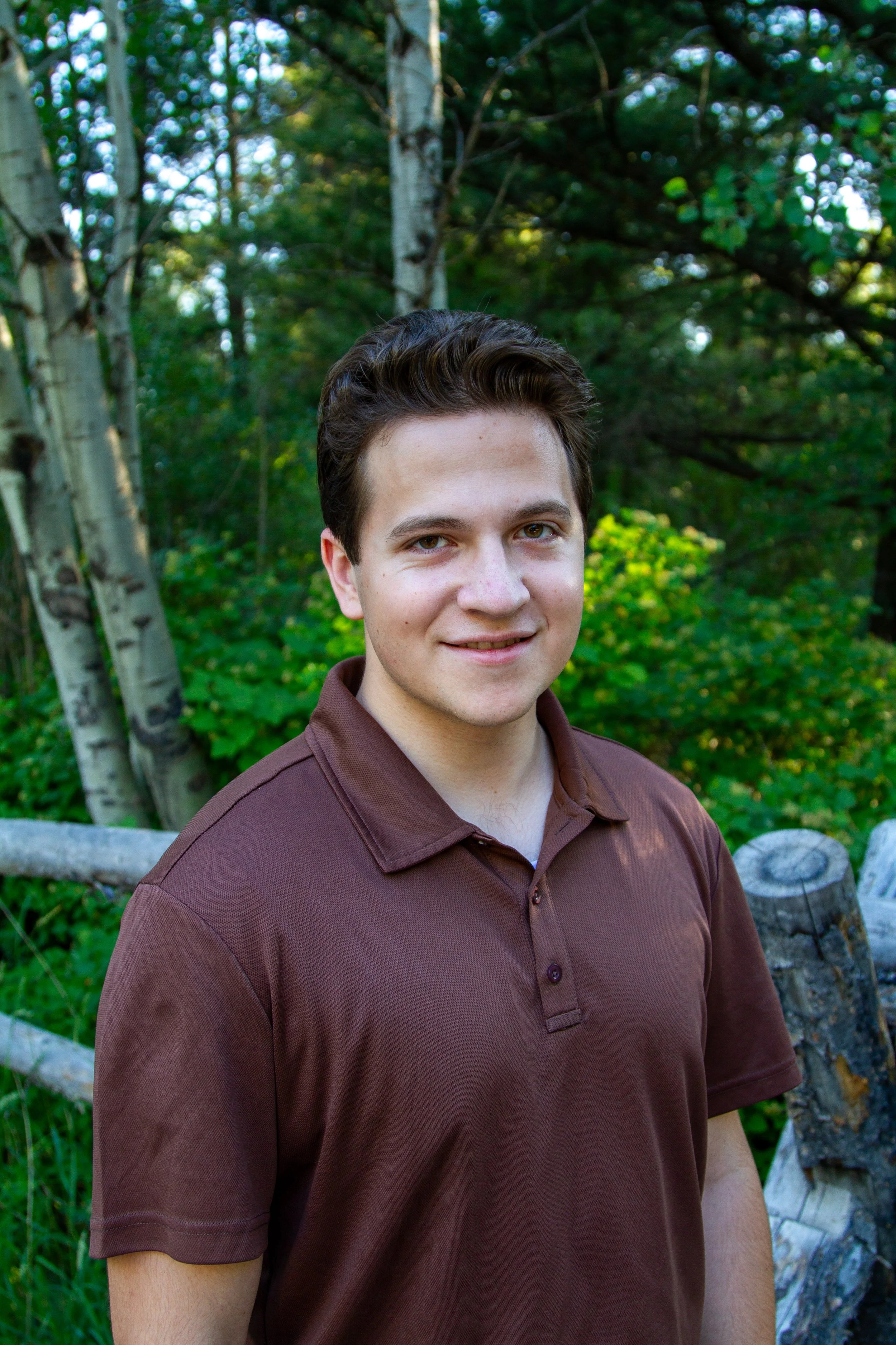 A young man with dark hair and light skin stands outdoors near trees and greenery, smiling at the camera.
