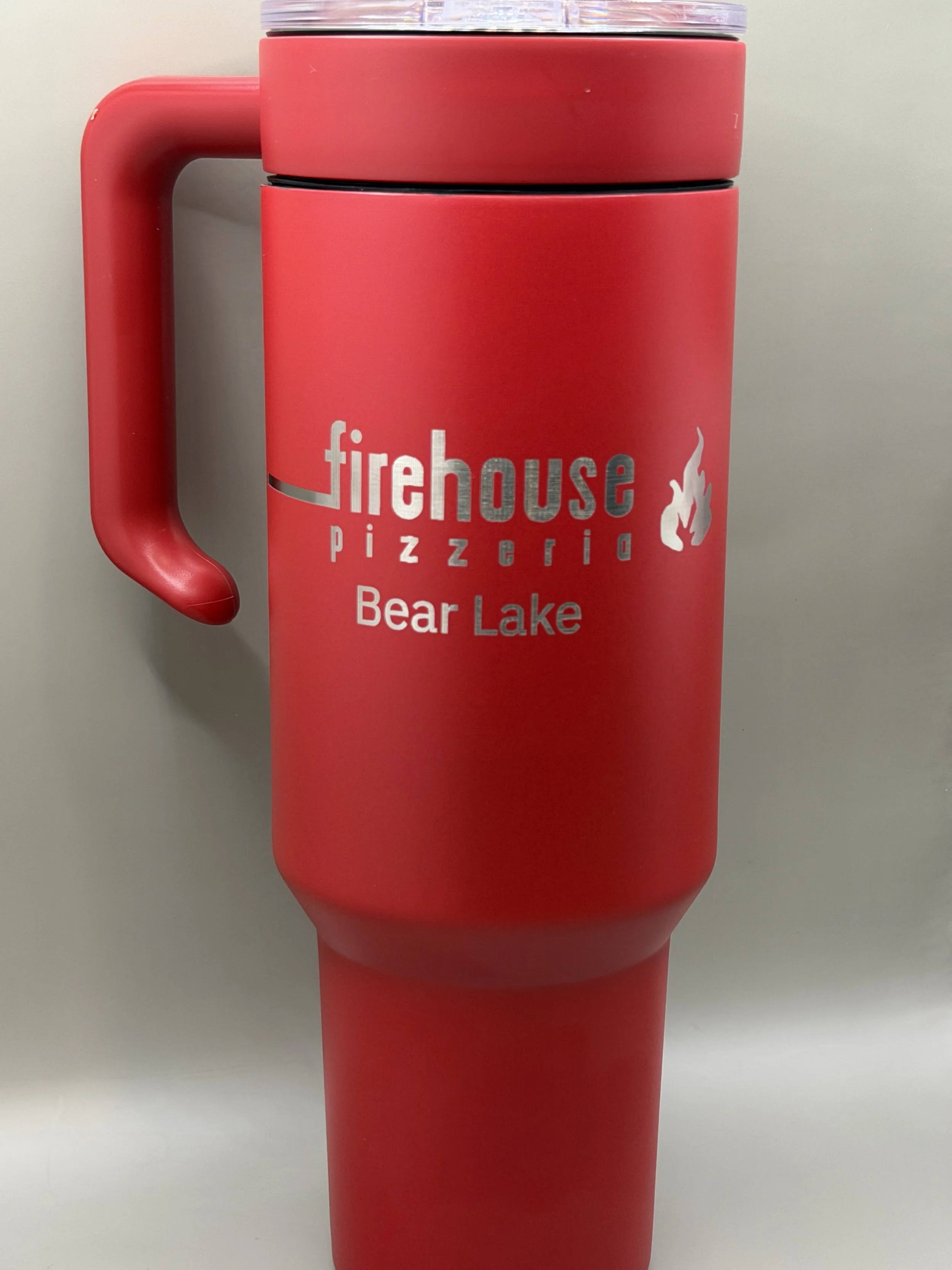 A red travel mug with a handle, with the logo in white and silver reading 'firehouse pizzeria Bear Lake' and a small flame icon, placed on a plain surface against a plain background.