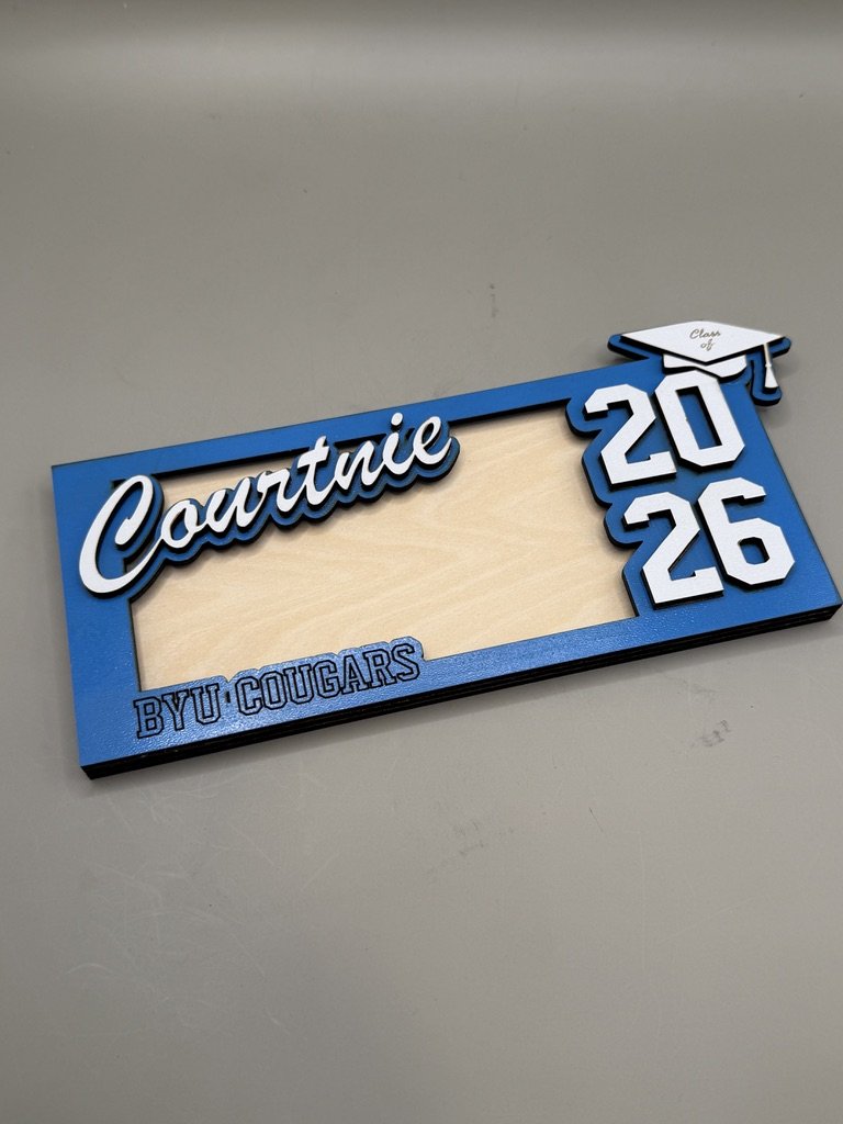 Blue graduation money holder in white cursive, the year "2026" in white block numbers, a small graduation cap at the top right corner, and the text "BYU COUGARS" at the bottom left.