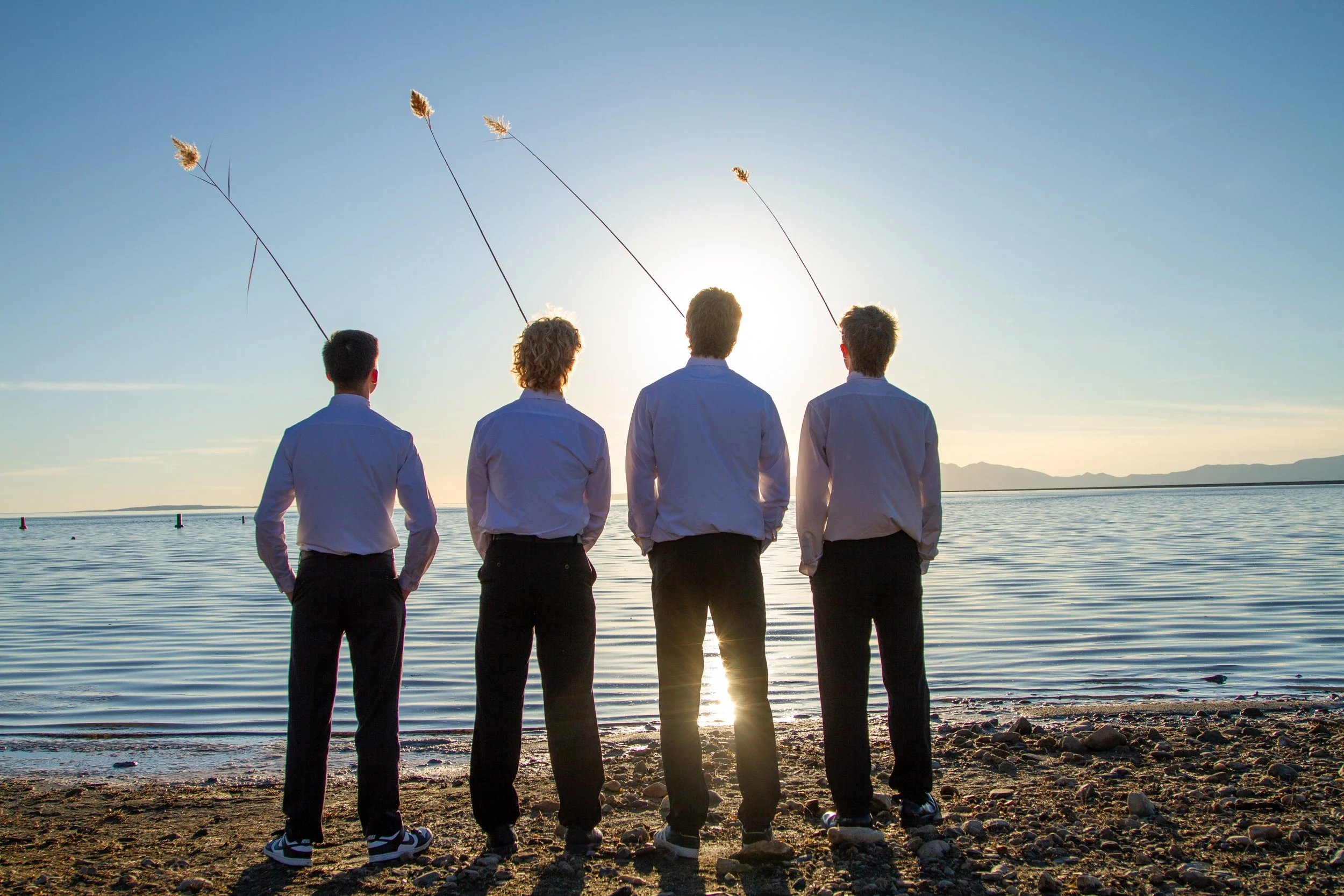 Four people standing on a rocky beach with their backs to the camera, facing the water with the sun setting behind them, one person holding three strings with wheat sheaves attached.
