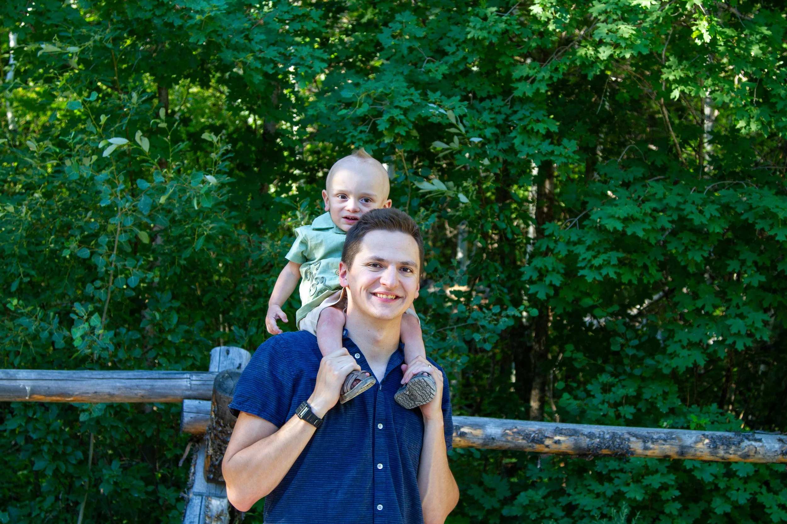 A man carrying a young child on his shoulders outdoors in front of green trees, smiling at the camera.