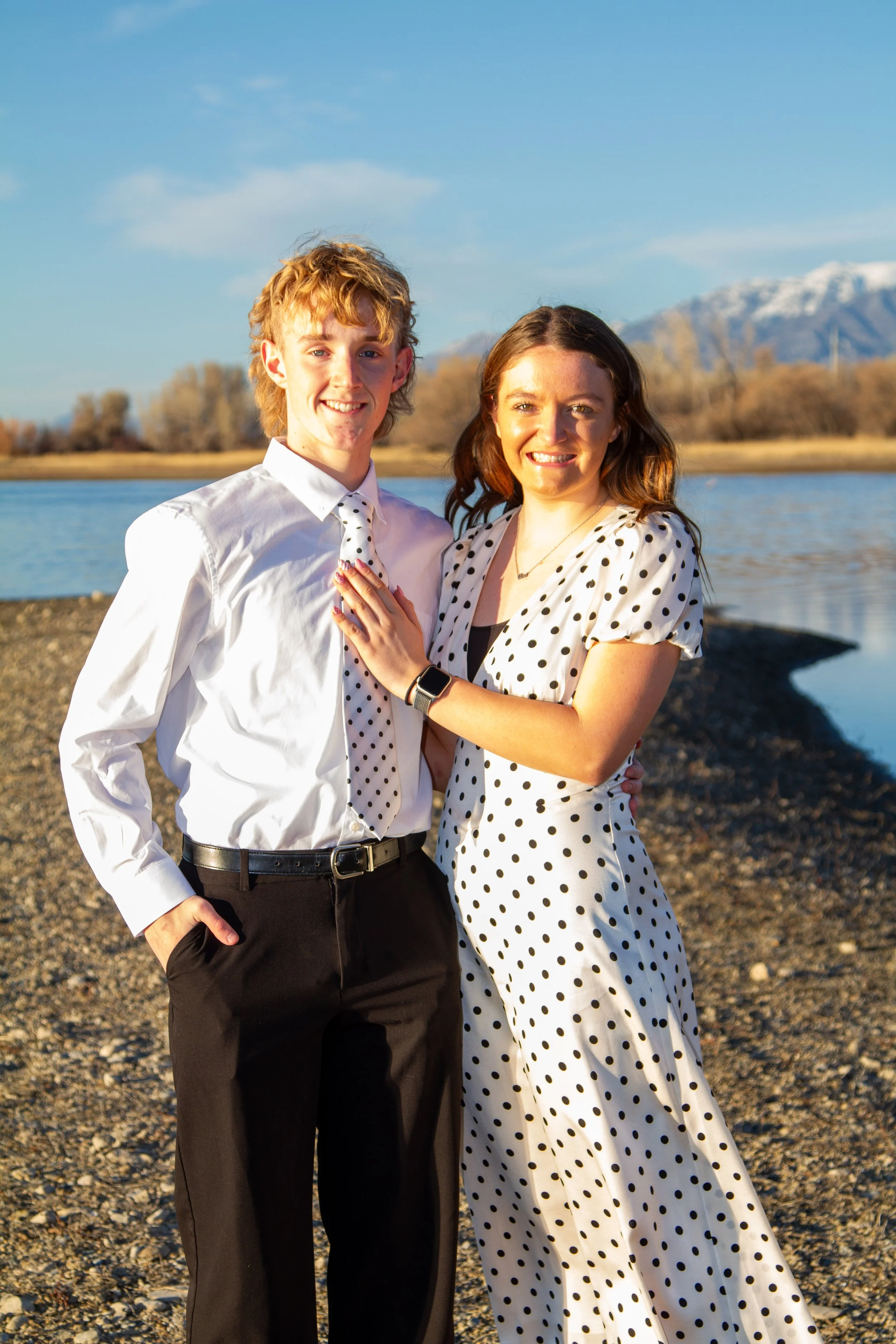 A young man and woman standing together near a body of water with mountain in the background during sunset, both smiling, dressed in white with black polka dots.