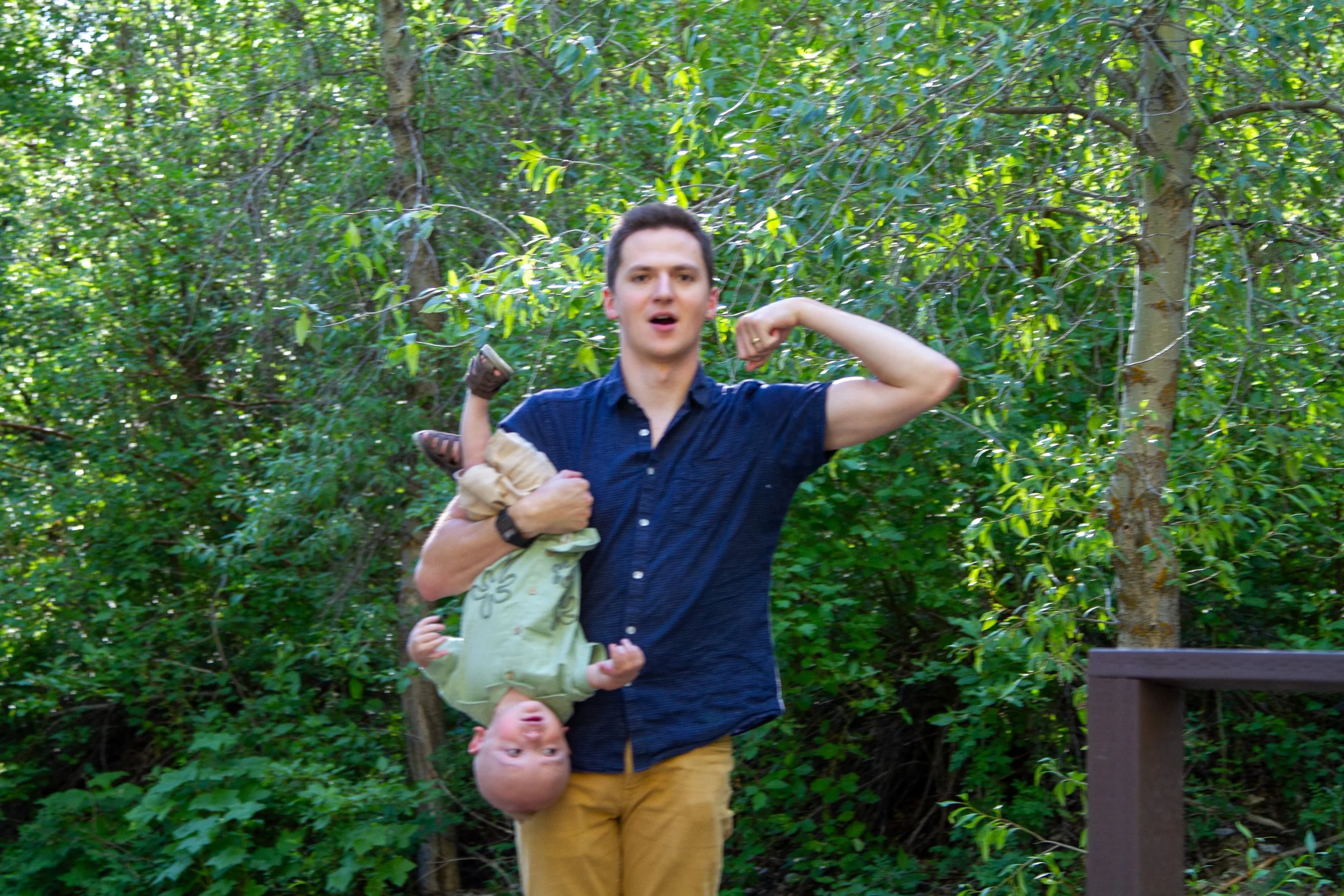 A man with short dark hair wearing a navy blue shirt and mustard yellow pants is carrying a young girl on his shoulders, who is upside down with her head hanging downward. They are outdoors in a wooded area with green leaves and trees.