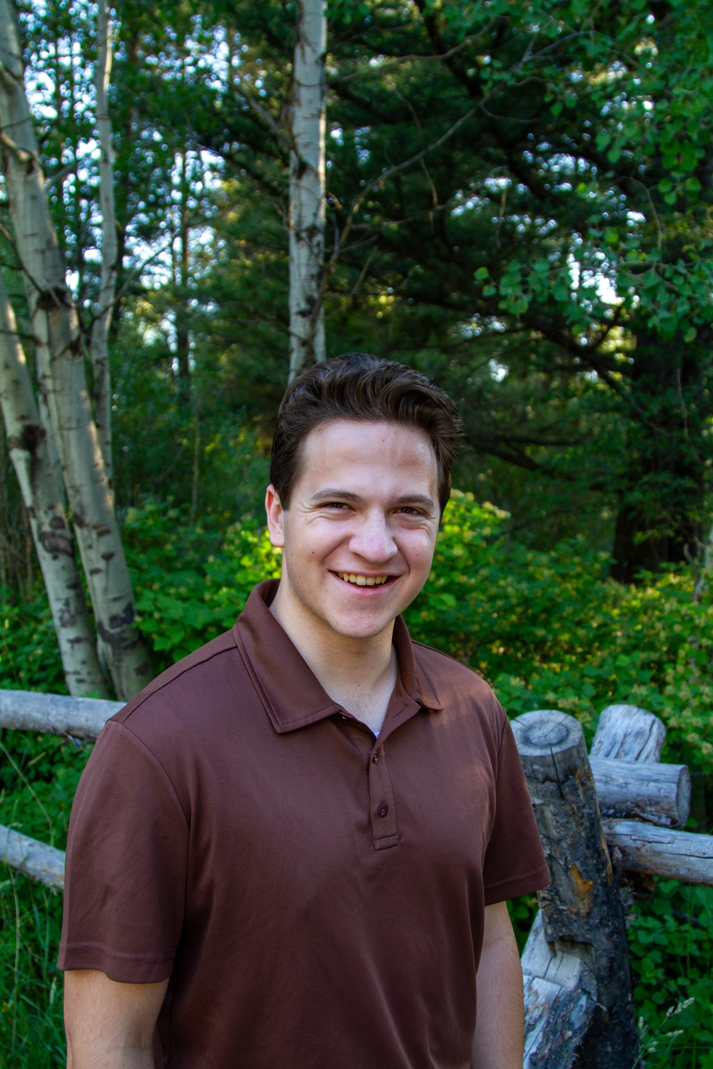 Young man with short brown hair smiling outdoors with trees, bushes, and fallen logs in the background.