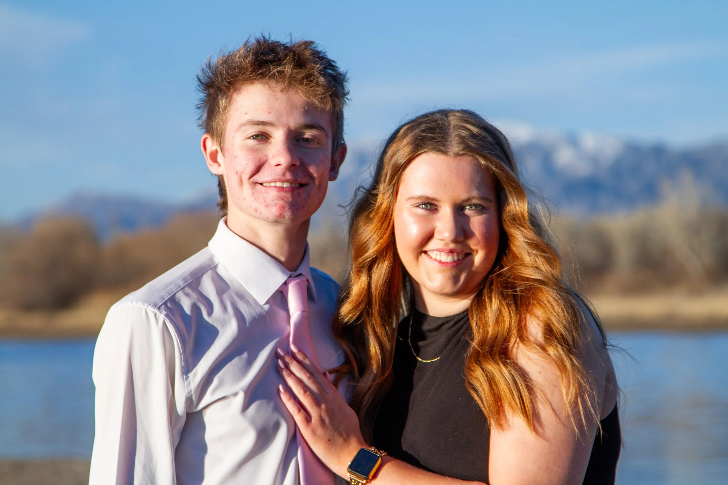 A young man and woman smiling, standing outdoors near a body of water with mountains and trees in the background.