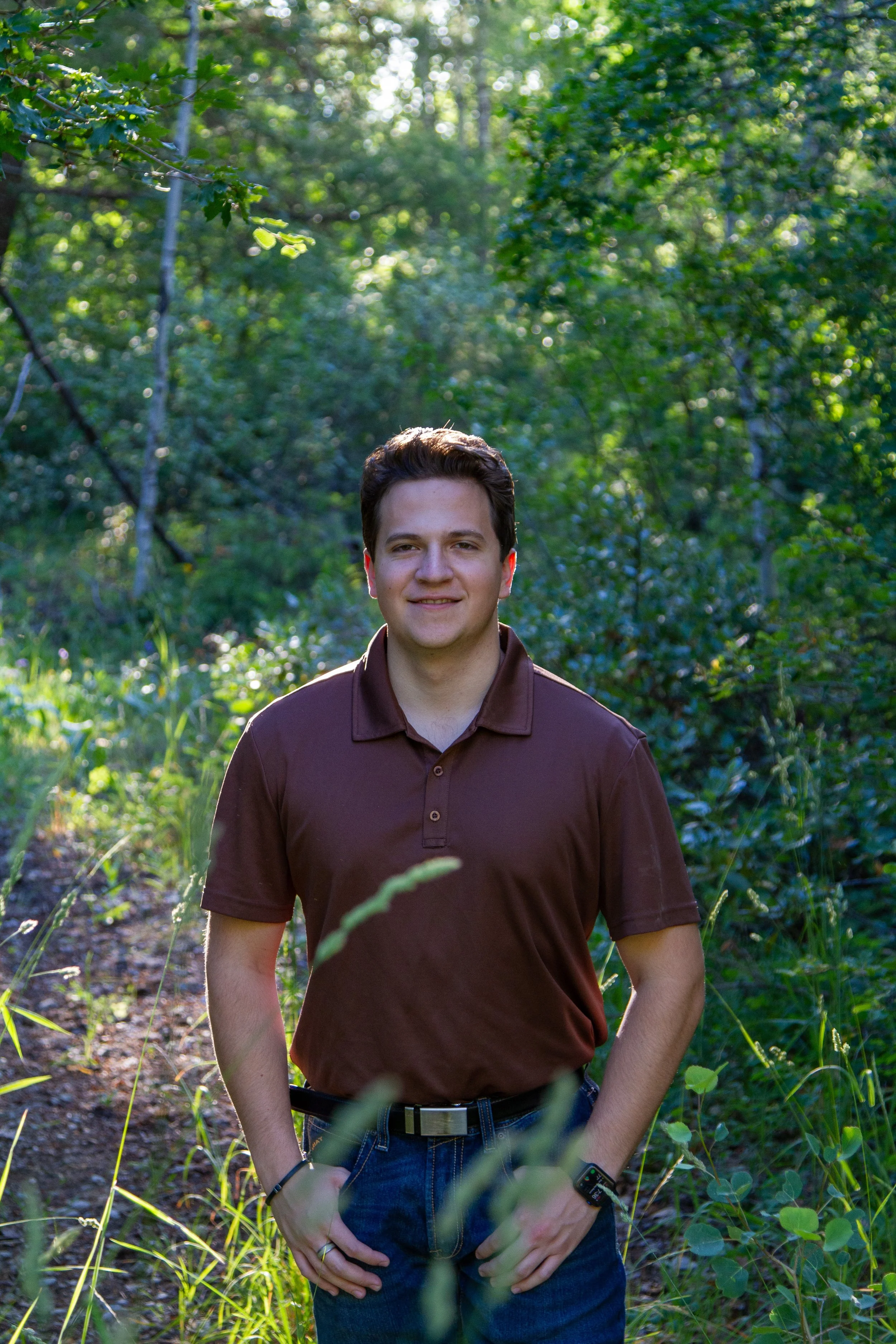 A young man with dark hair smiling outdoors in a green wooded area, wearing a dark brown polo shirt and jeans.