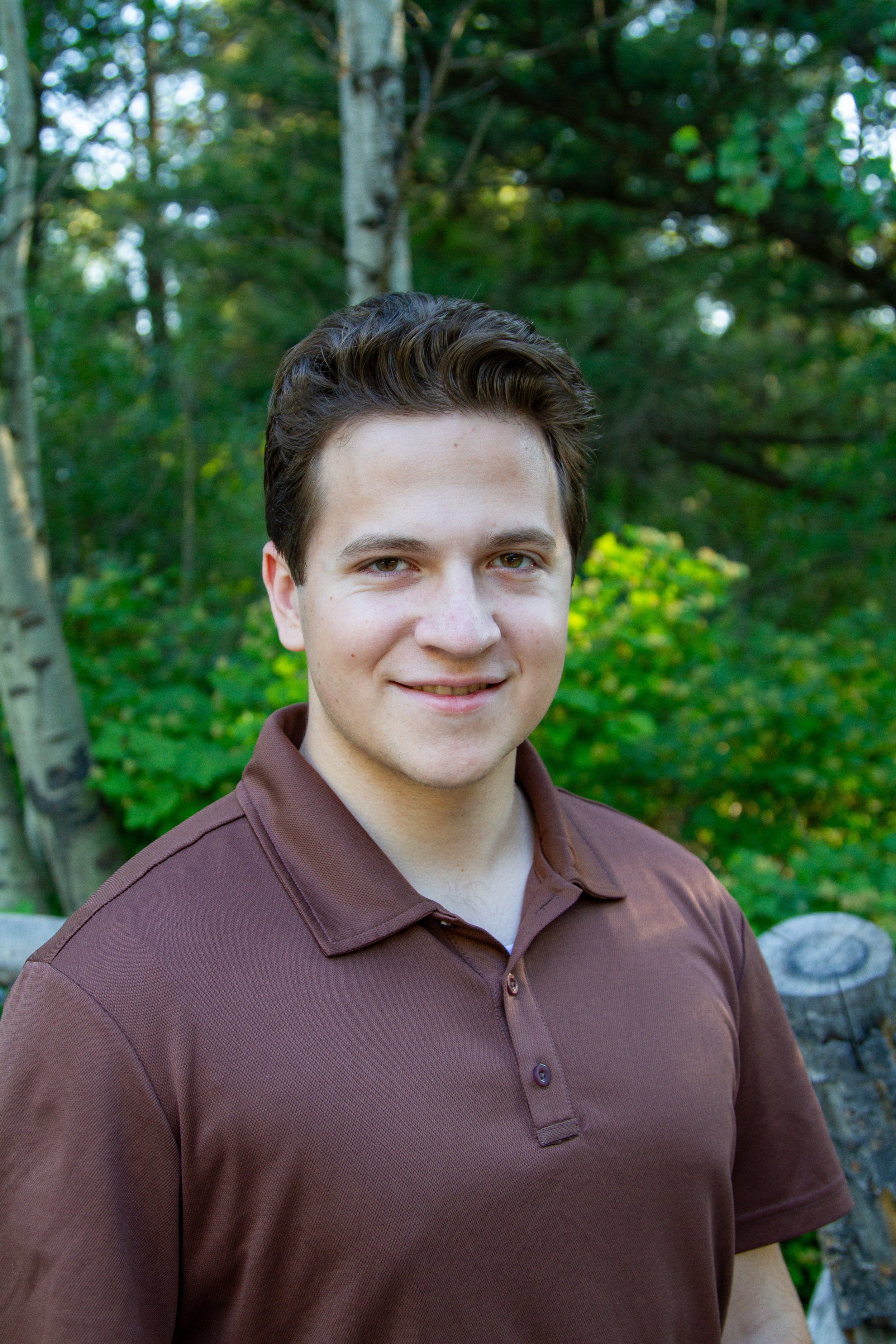 A young man with light skin, short brown hair, and a friendly smile, wearing a brown polo shirt, standing outdoors in a wooded area with green trees and rocks in the background.