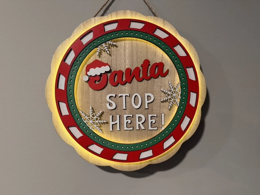 Decorative Christmas sign with the words "Santa STOP HERE!" in red and white lettering, surrounded by a red and green border, with snowflakes and a Santa hat on the word "Santa."
