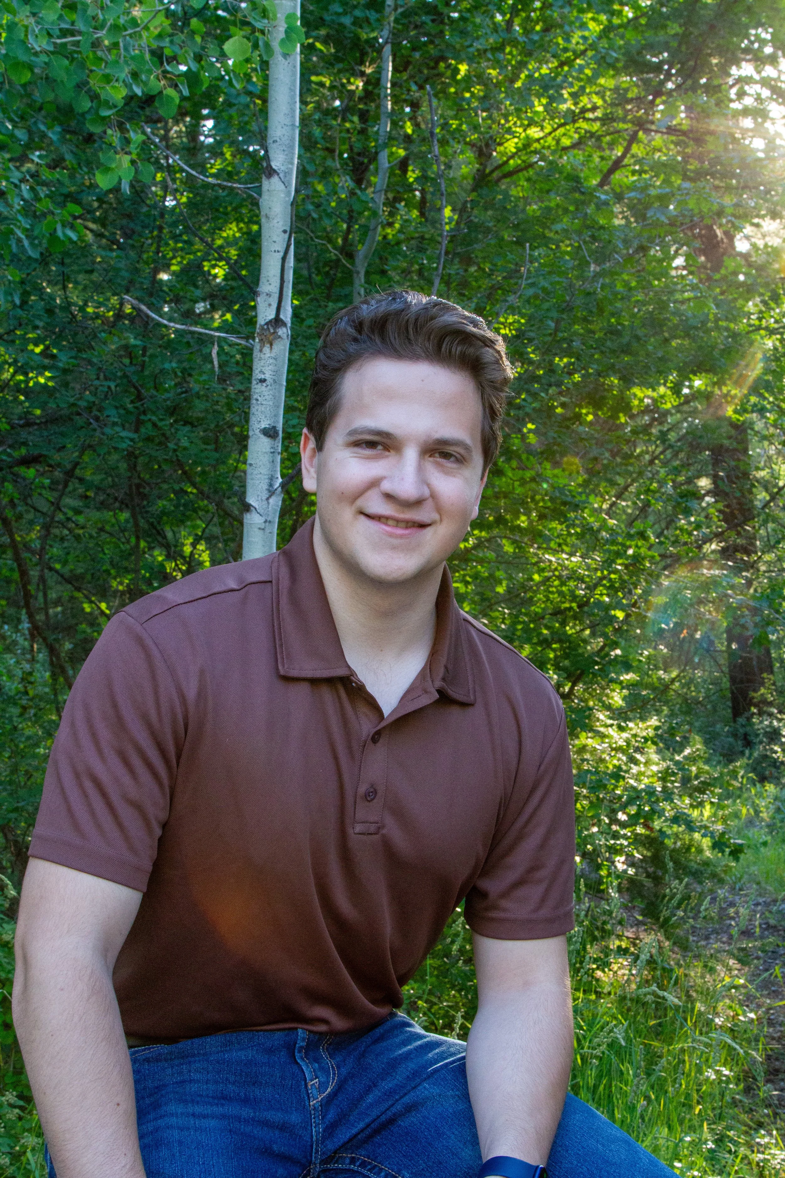 A young man with dark hair, wearing a maroon shirt and blue jeans, sitting outdoors in a wooded area with green trees and sunlight in the background, smiling at the camera.