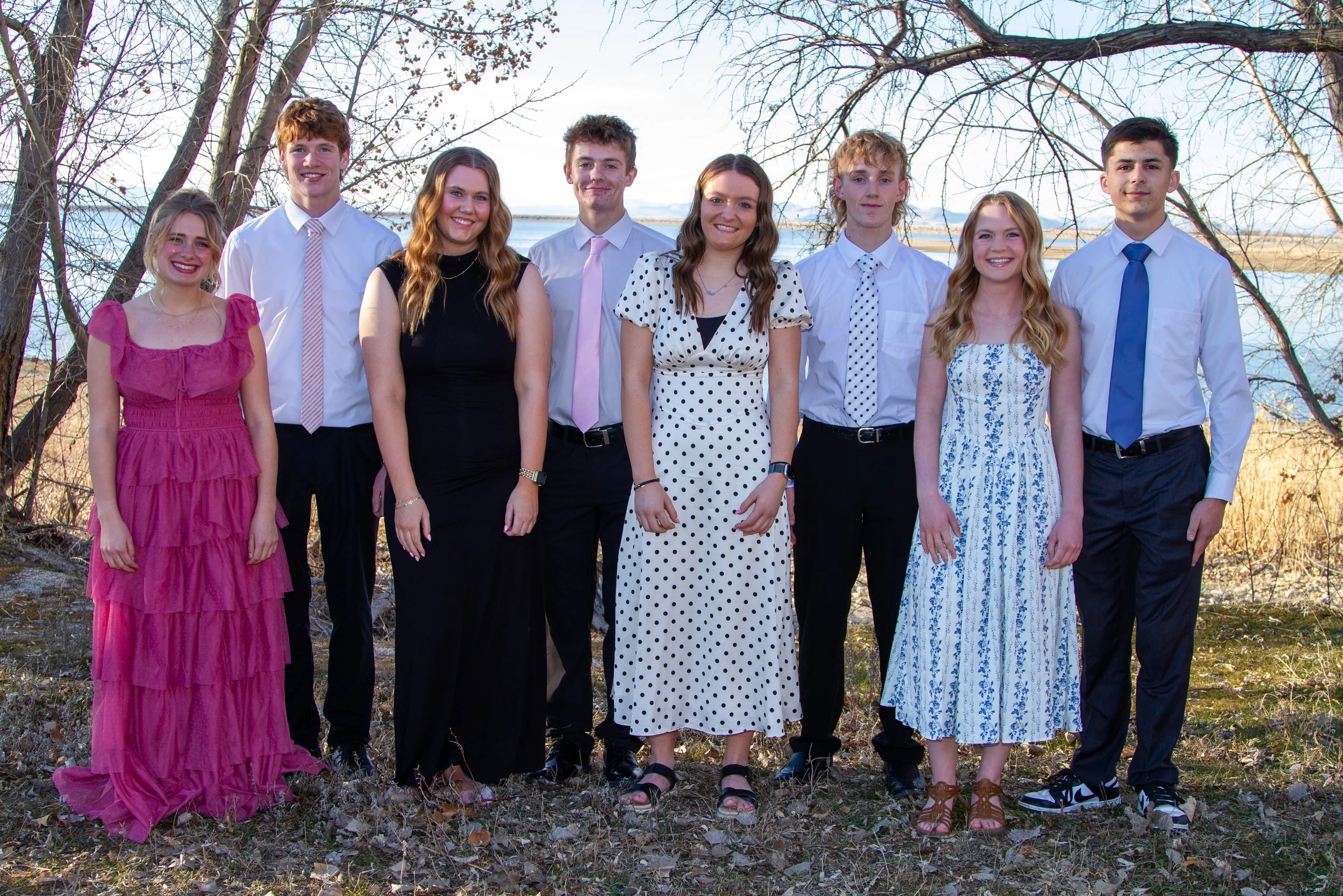 Group of nine young people outdoors near a body of water, standing on grass and leaves, dressed in semi-formal attire, with trees in the background.