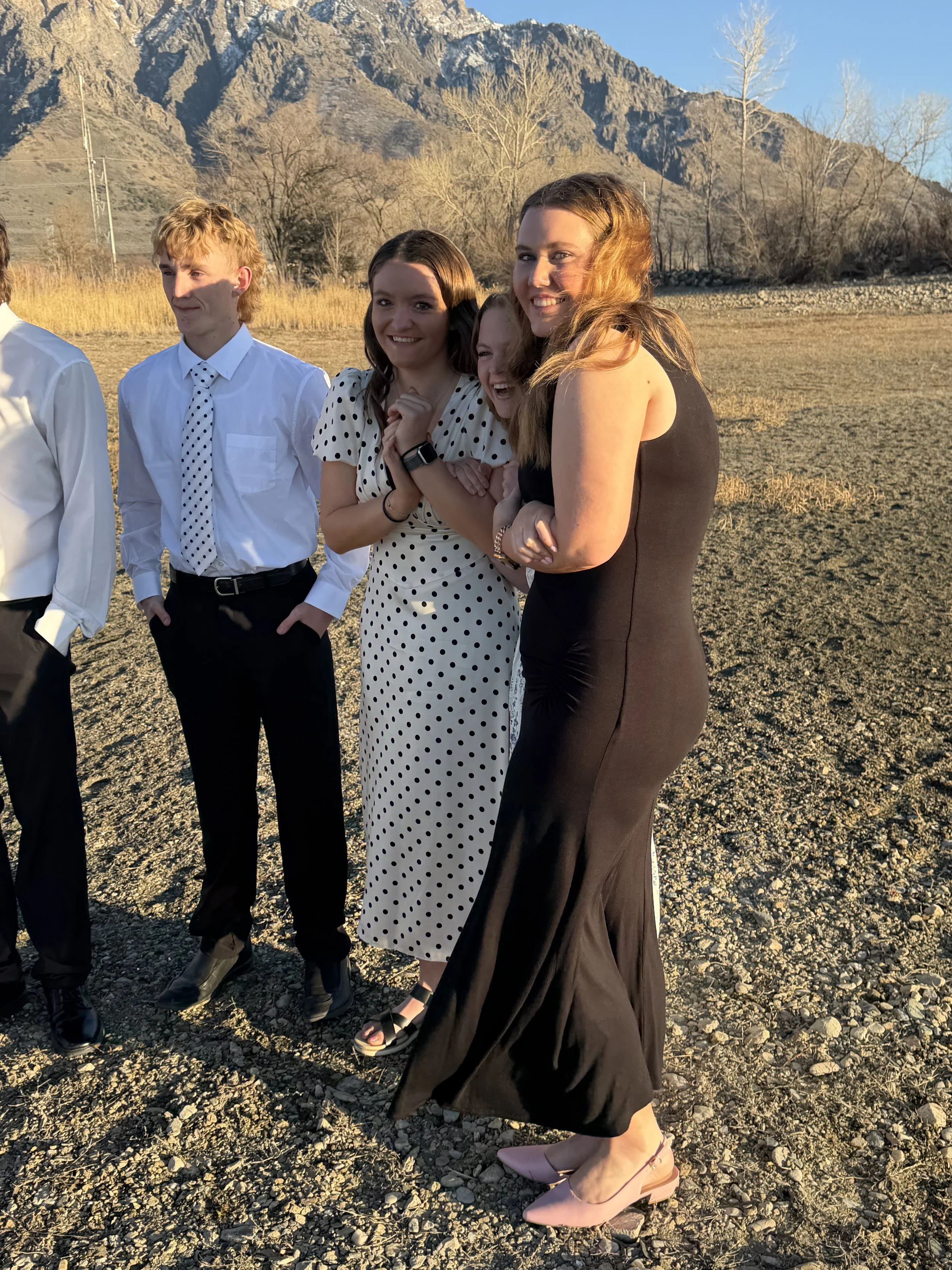 A group of four young women and one young man standing outdoors on rocky terrain, with mountains and sparse trees in the background, dressed in semi-formal attire, smiling and enjoying the sunny weather.