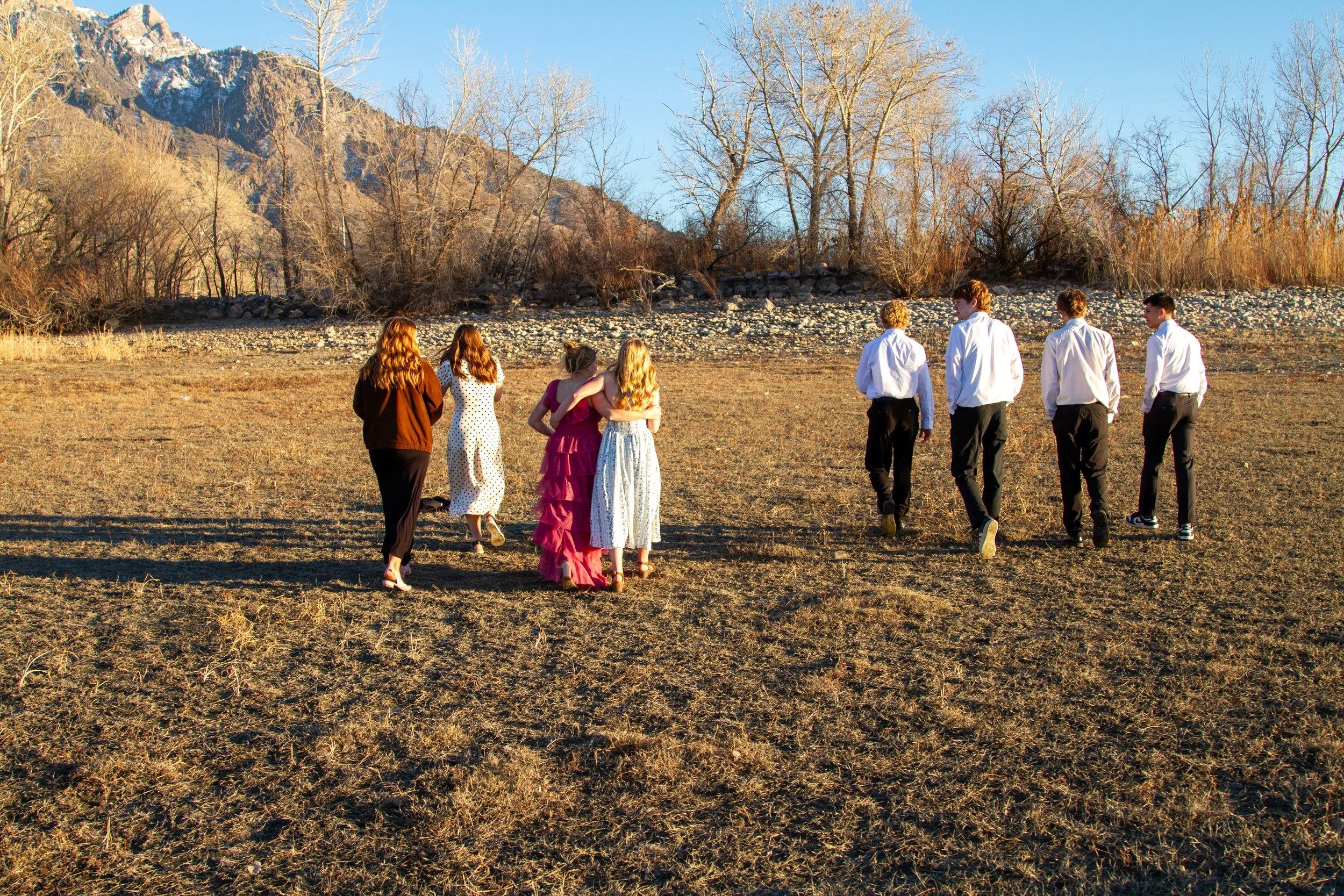 A group of seven young people walking across a dry, grassy field with mountains and bare trees in the background during late afternoon or early evening.