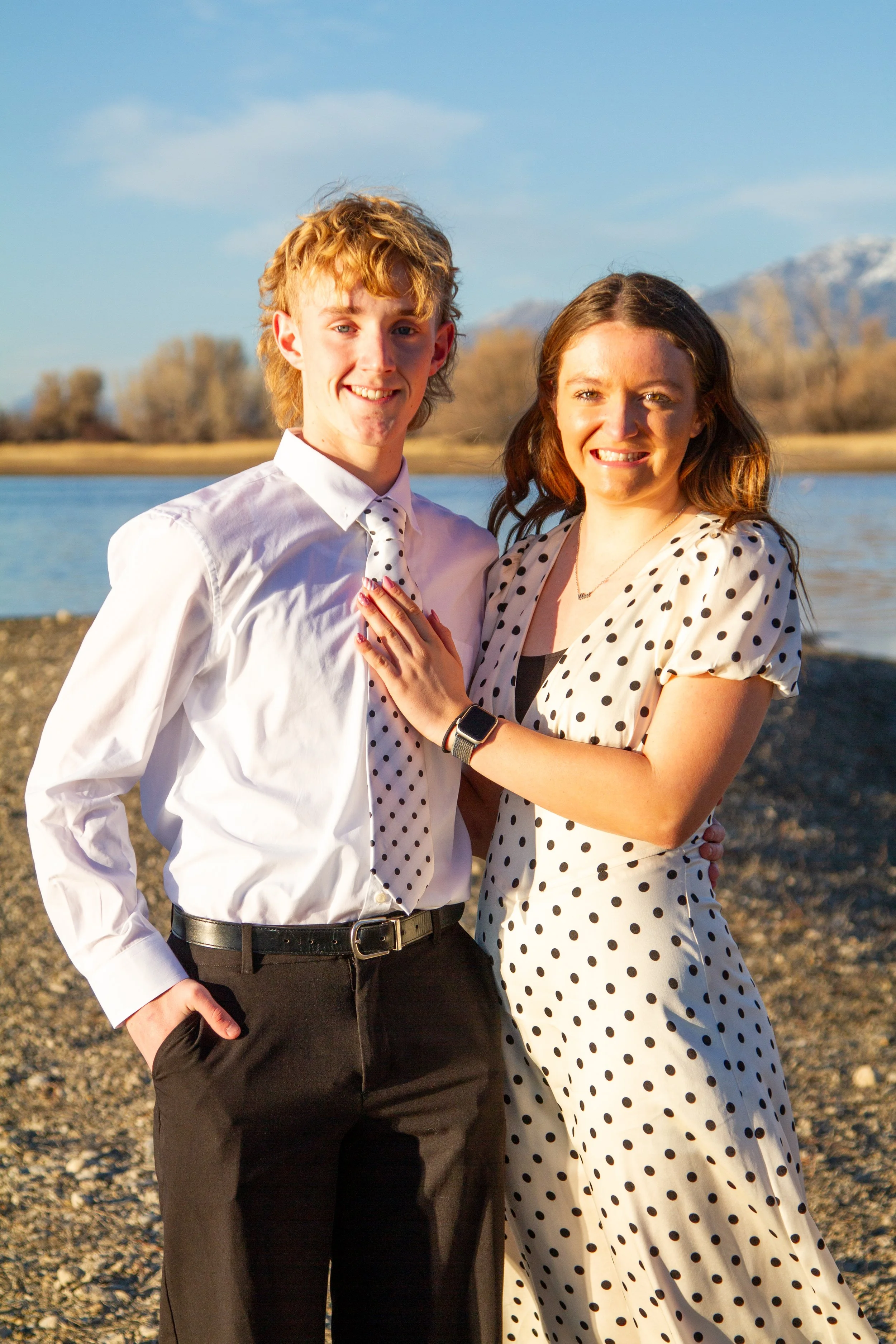 A young man and woman standing outdoors near a body of water, smiling at the camera. The man is wearing a white shirt and black pants, and the woman is wearing a polka dot dress. The background includes trees and mountains.