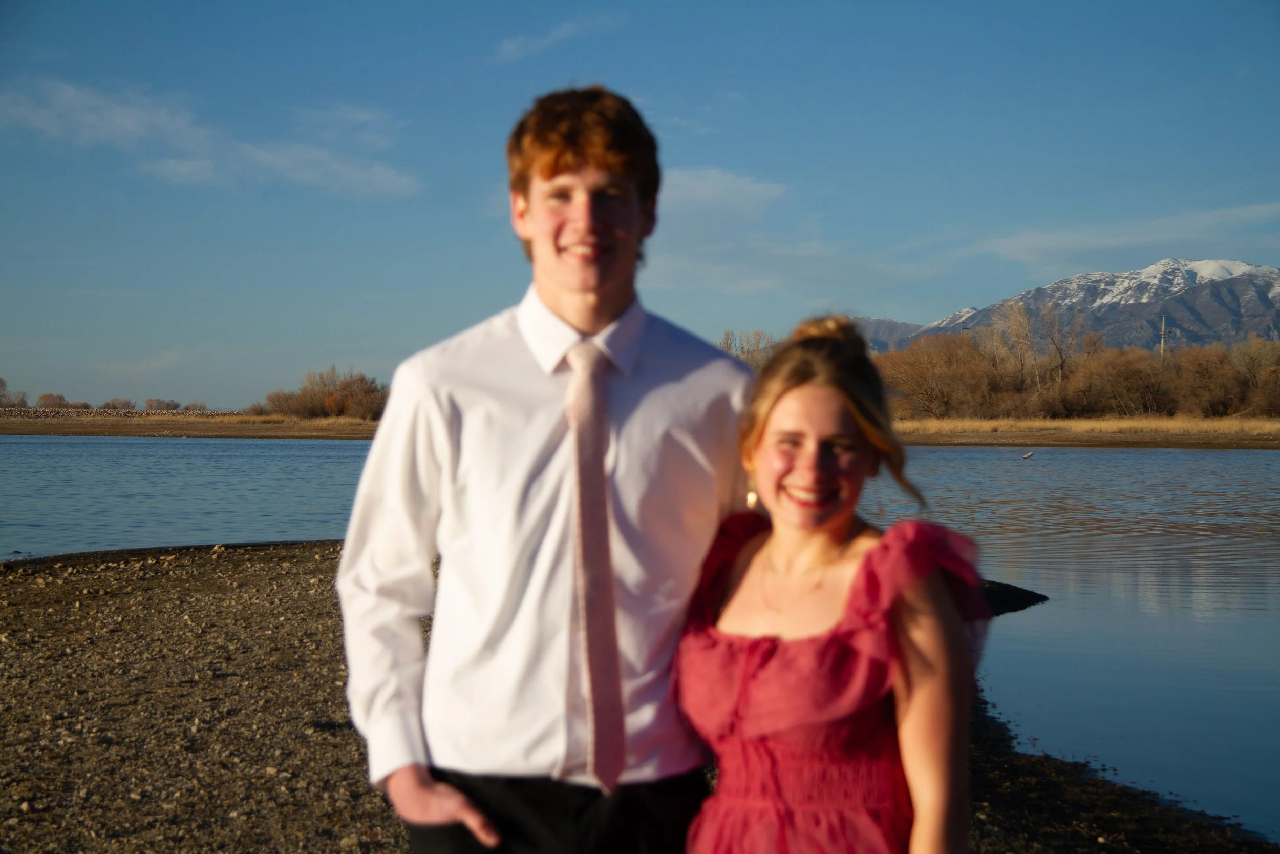 A smiling man and woman standing by a body of water with mountains in the background, during sunset or late afternoon.