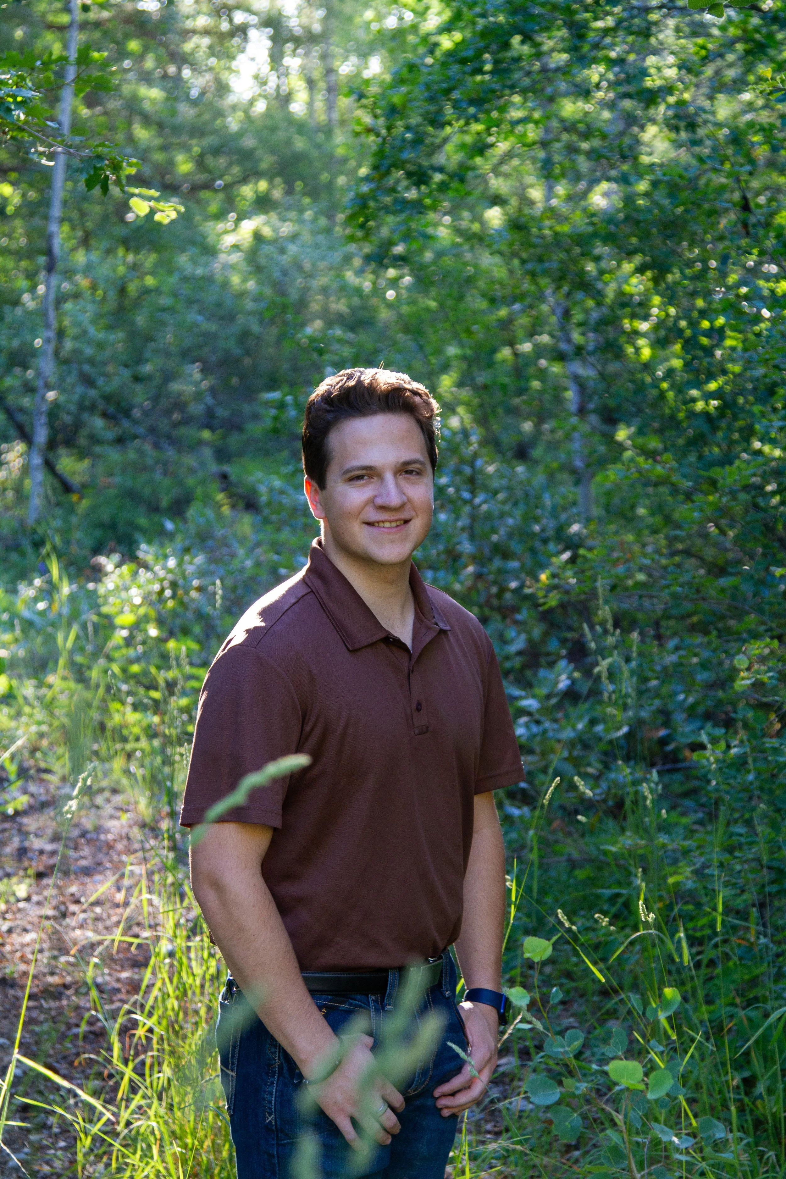 A young man in a brown polo shirt standing outdoors in a lush green forest with sunlight filtering through the trees.