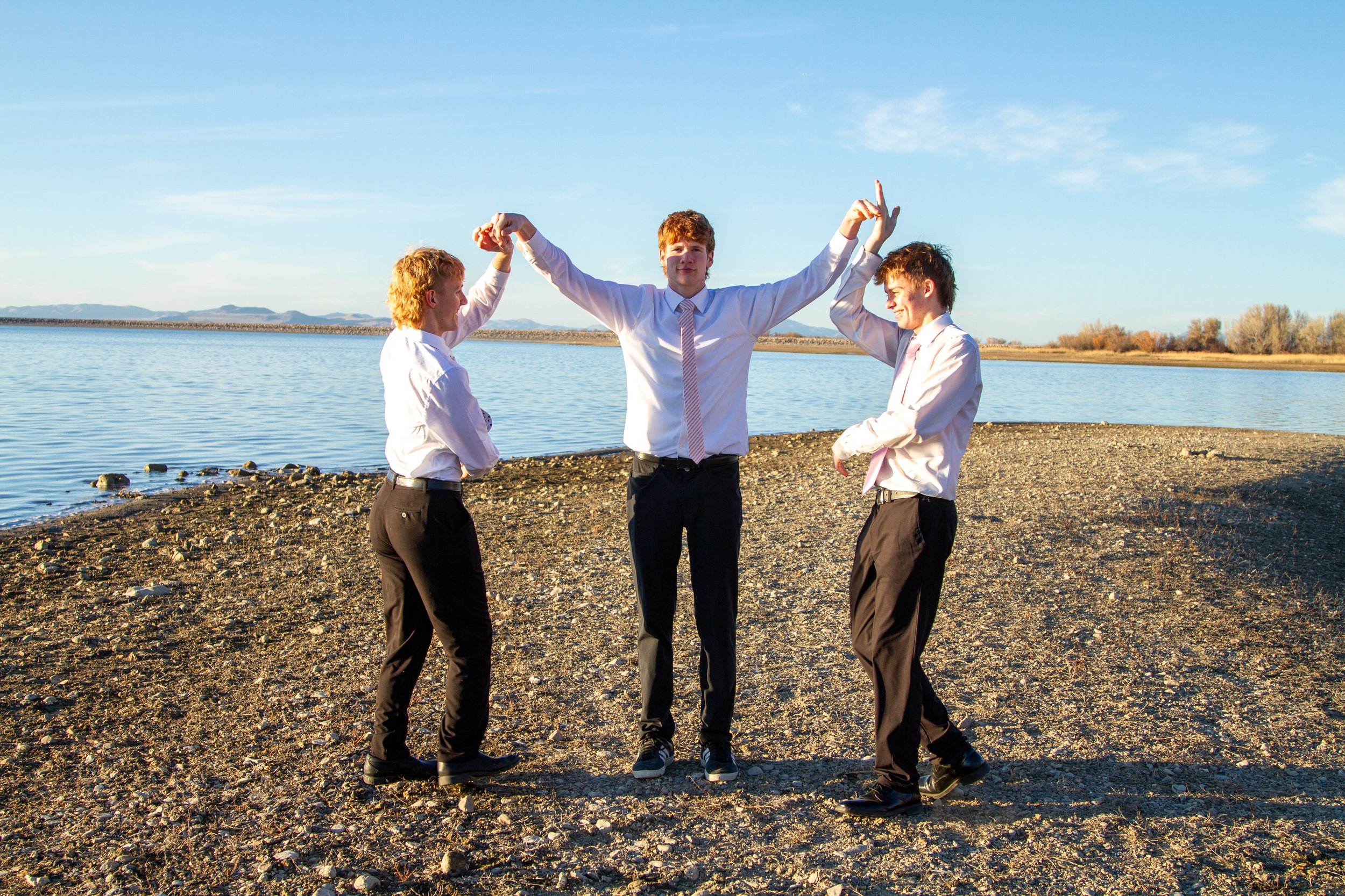 Three young men dressed in business casual, standing on a pebbly shoreline near a body of water, with mountains in the background. The man in the center, with red hair, raises his arms as the other two hold his hands.