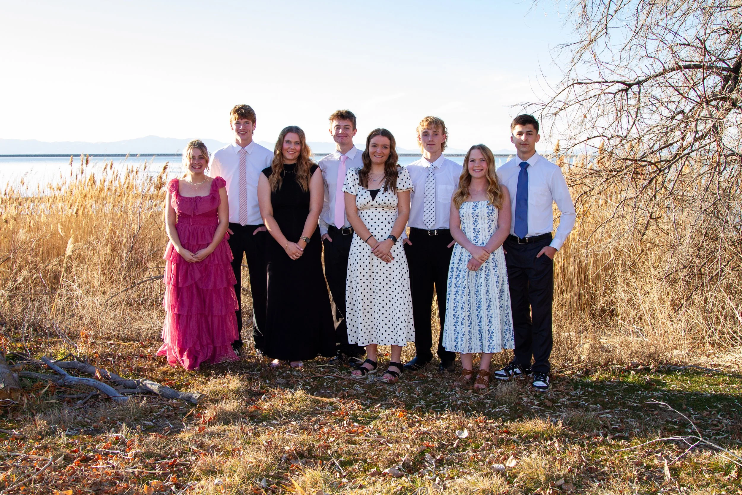 Group of nine teenagers standing outdoors on grass with tall dry grass and trees in the background, dressed in semi-formal attire.