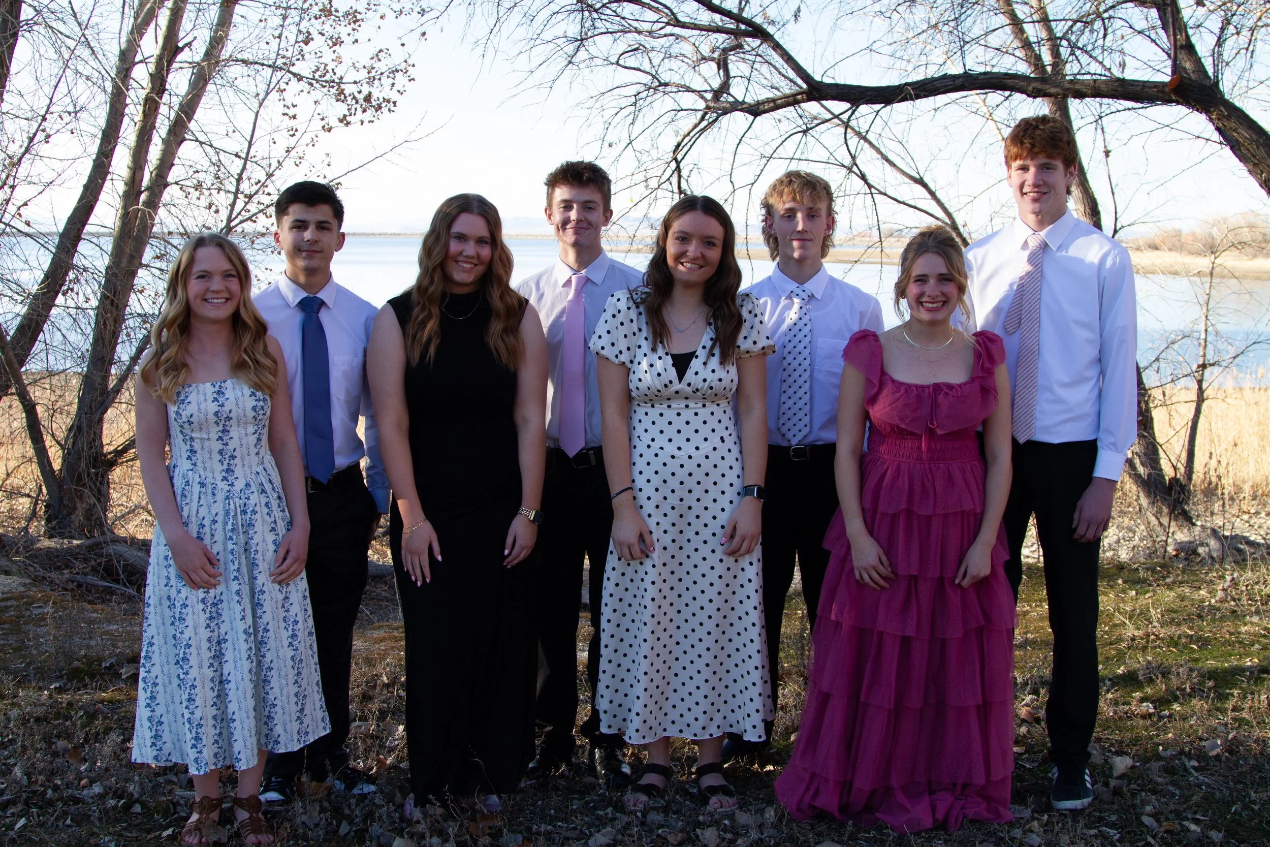 Group of eight young people standing outdoors near a body of water with trees in the background, dressed in semi-formal attire, smiling for a photo.