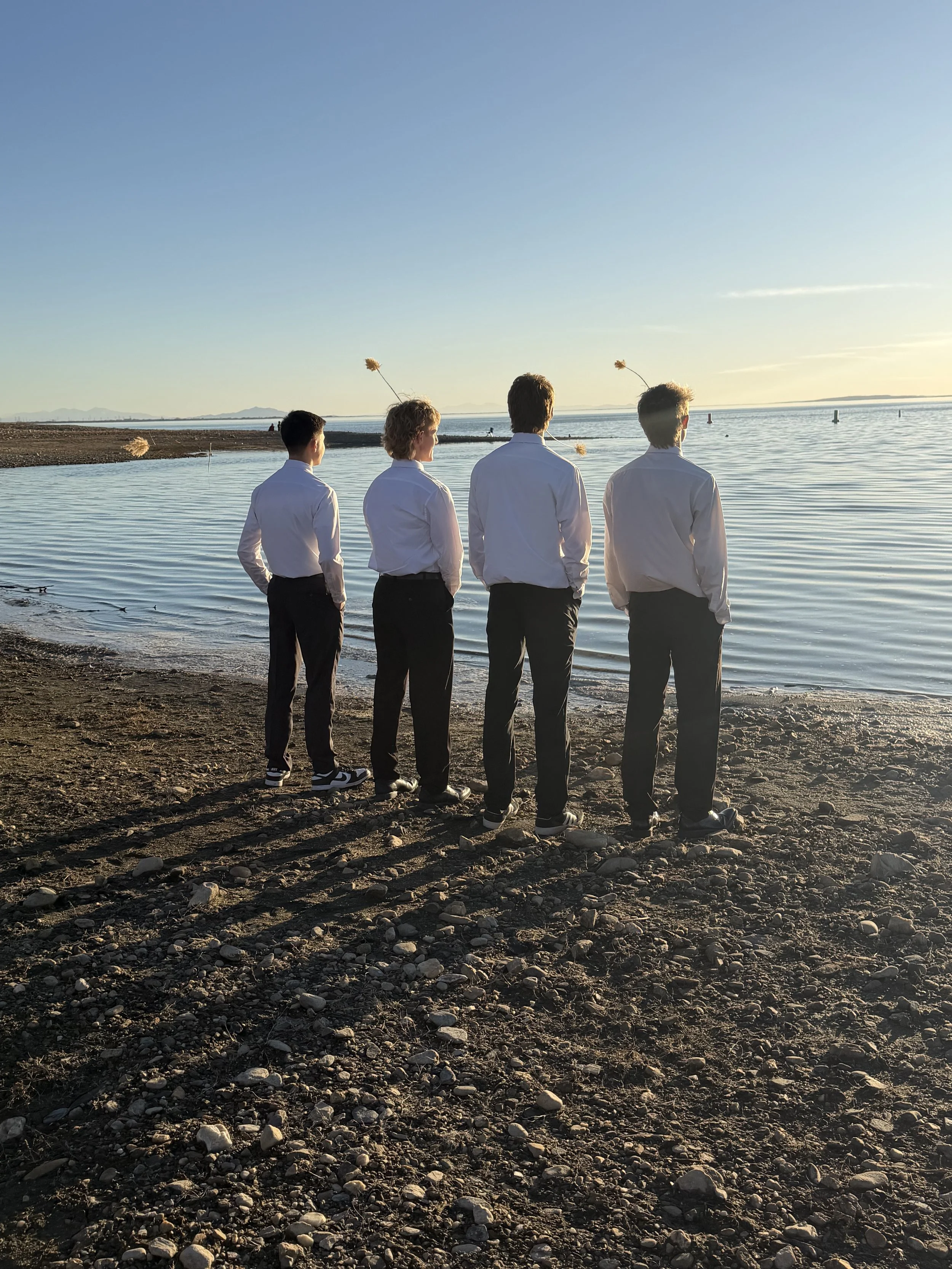 Four men in white shirts and dark pants standing on a rocky beach facing the water during sunset.