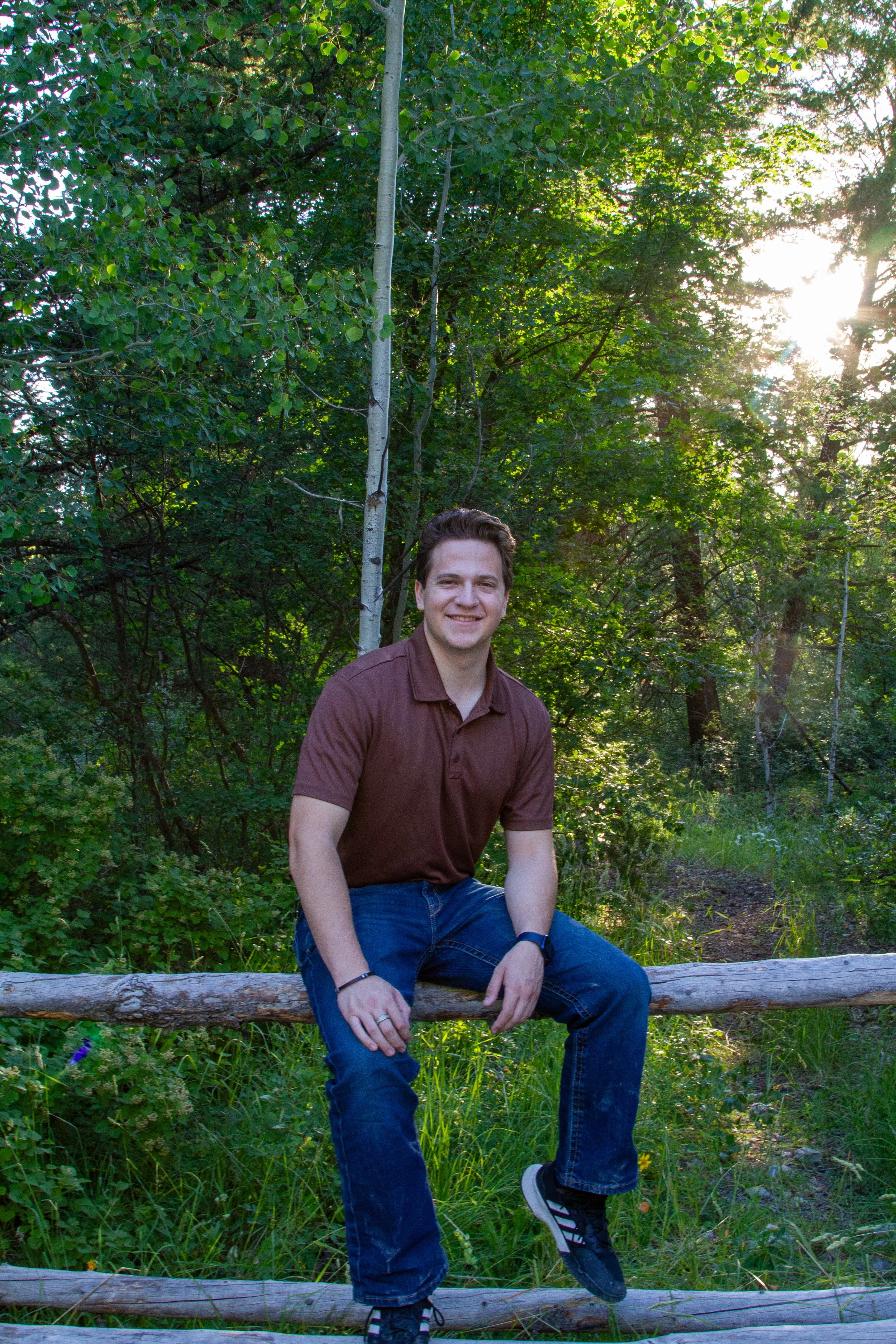 Young man sitting on a wooden log in a forested area, smiling, wearing a maroon polo shirt and jeans, with sunlight filtering through the trees.